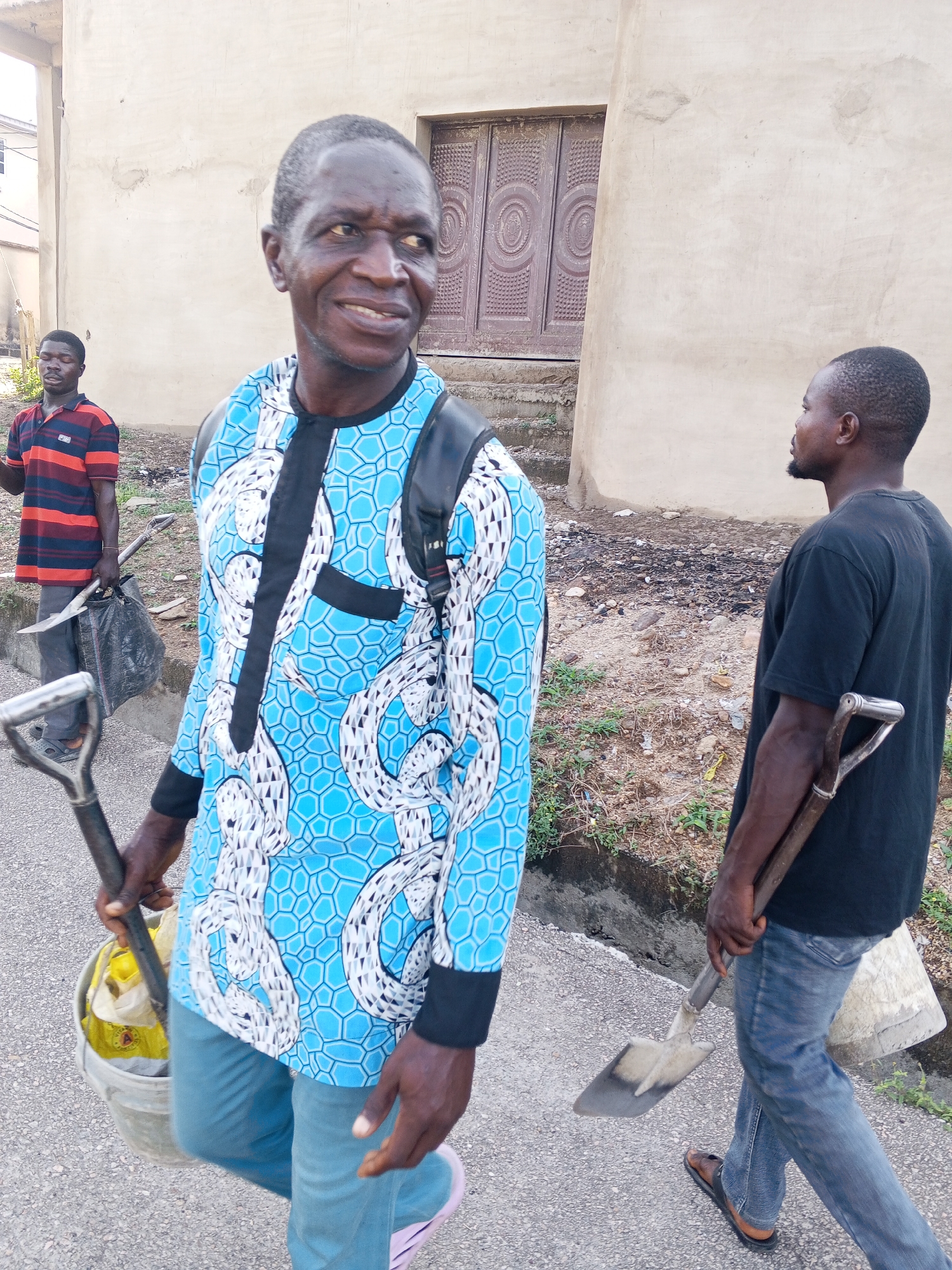 The image depicts three men on a street in Lokoja, Nigeria. The man in the foreground is wearing a blue, long-sleeved shirt with a white snake-like pattern, black trim, and blue jeans. He carries a bucket and shovel. A second man in the middle ground wears a black t-shirt and blue jeans and carries a shovel over his shoulder. The third man is further in the background, wearing a horizontally striped shirt and carrying a black plastic bag. In the background is a beige building with brown doors. The ground is paved, with vegetation and what appears to be discarded waste along the side.