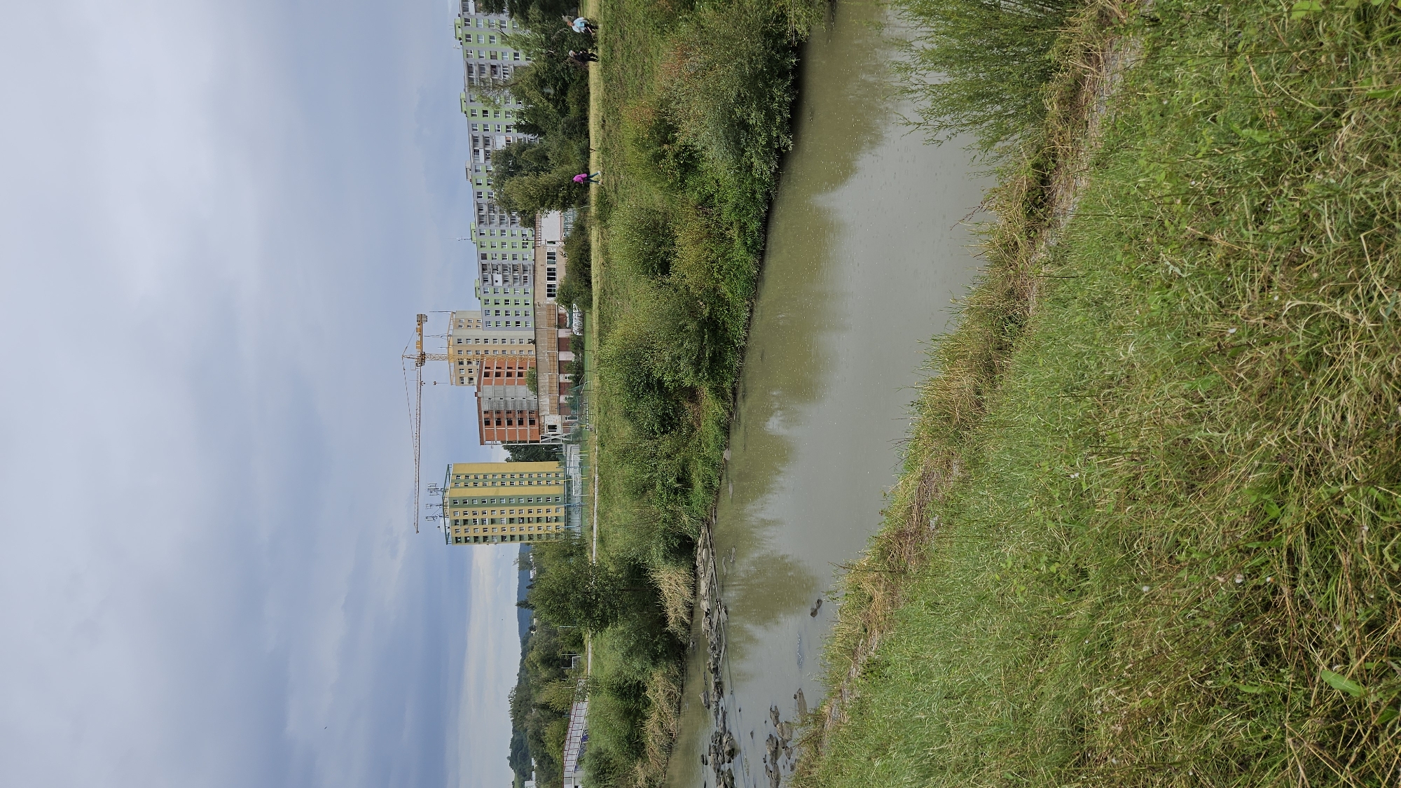 The image captures a view of the Sekčovský stream in Prešov, Slovakia, on a cloudy day. In the background, a series of apartment buildings line the horizon, with construction cranes visible near two of the taller structures, one yellow and one peach-colored, suggesting ongoing development. A lower, light brown building is situated between the construction sites. The stream, with murky water and scattered rocks, flows across the foreground, bordered by grassy banks and dense green bushes. A white footbridge arches over the stream on the left side. Several people are walking along the grassy area beside the stream on the right.
