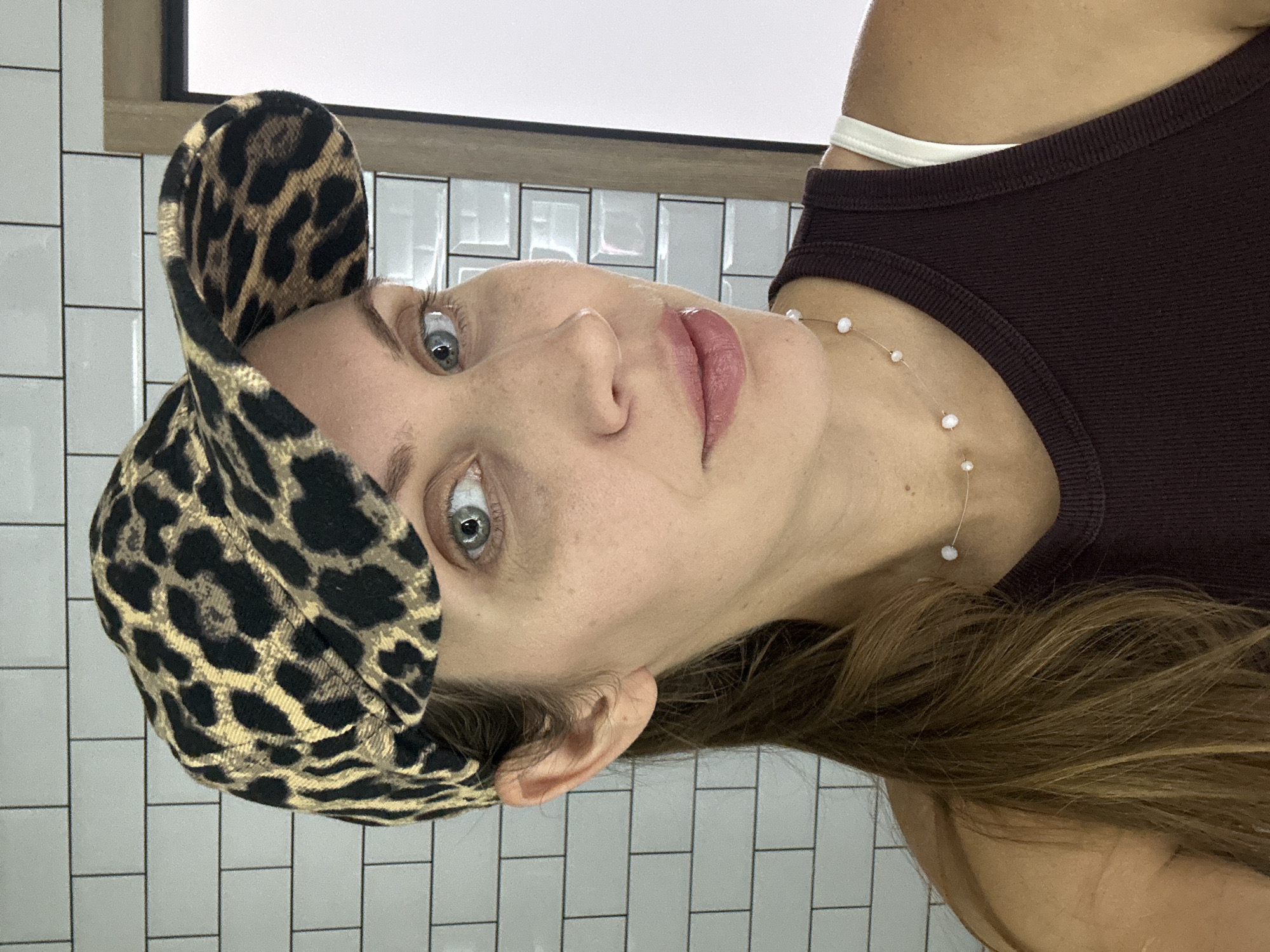 A woman with long brown hair, wearing a leopard print baseball cap and a dark brown tank top with a white bra strap visible, poses for a selfie. She has blue eyes, light skin with freckles, and is wearing a delicate necklace with small white beads. The background features white subway tiles. The image was taken in a room with a mirror to the right.