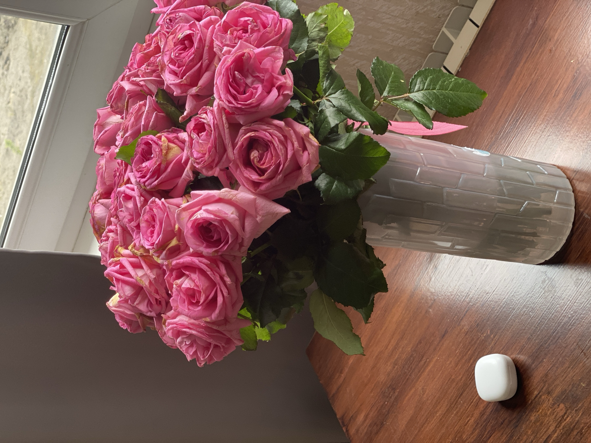 A still life photograph shows a bouquet of pink roses in a grey vase on a wooden table. A small, white, rounded square object is also present on the table near the vase. The setting is indoors, likely near a window as indicated by the light source on the left. There is no specific information to indicate that this scene is in Sochi, Russia.