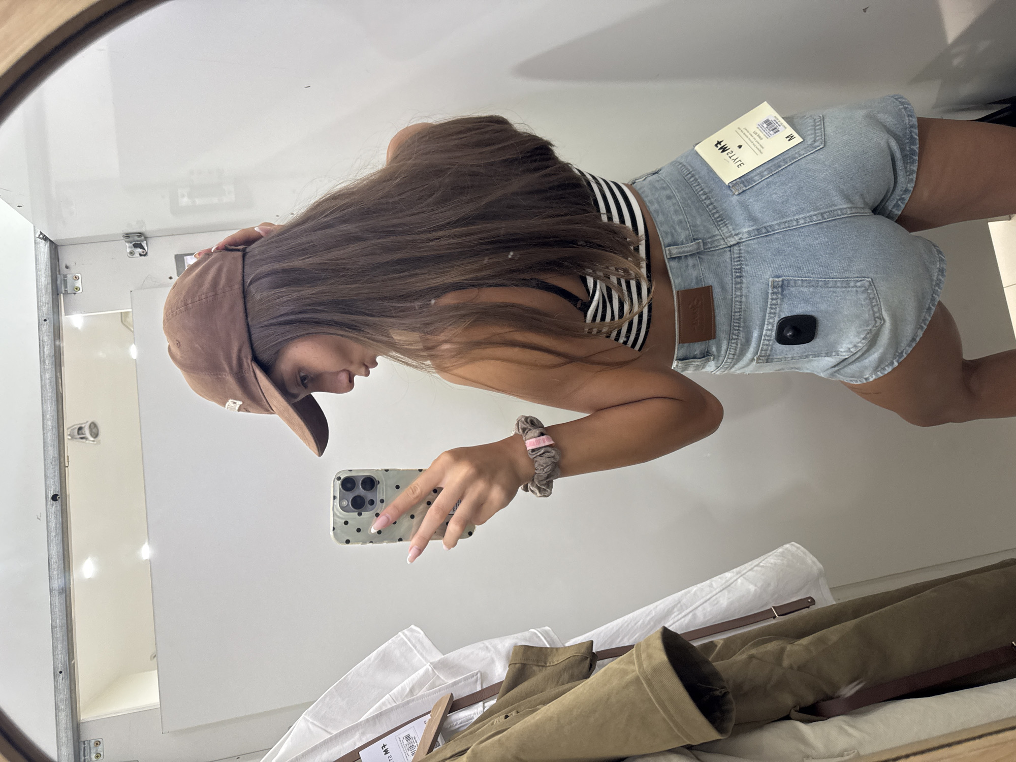 A young woman stands in a dressing room, visible in a mirror's reflection. She is wearing a brown baseball cap, a striped crop top, and light blue denim shorts with a tag still attached. She holds a smartphone with a polka-dot case, taking a selfie. Below her, clothes hang on a rack, including a white shirt and an olive green jacket. The dressing room is white and appears clean. The location is not discernible from the image.