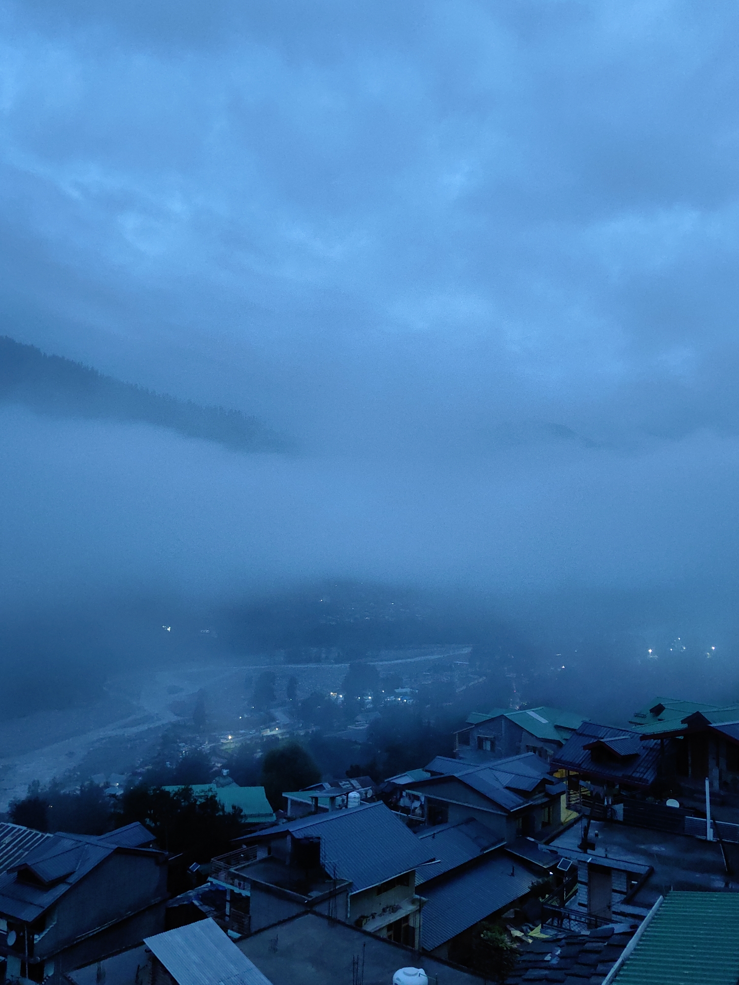 The image depicts a view of Vashisht, India, at dusk or dawn, characterized by heavy fog and low light conditions. The foreground features a dense cluster of multi-story buildings with predominantly sloped, dark-colored roofs, some of which are metallic. Several structures appear to be residential homes, with visible windows, some illuminated from within, suggesting occupancy. The architecture is varied, with some buildings exhibiting simpler designs and others appearing more elaborate.
The middle ground shows a village or town situated on a hillside, partially obscured by fog. Scattered lights are visible throughout this area, indicating the presence of more dwellings and infrastructure. The fog creates a hazy atmosphere, softening the details of the landscape and creating a sense of depth and mystery.
In the background, a mountainous terrain is faintly discernible through the fog. The overall color palette is dominated by shades of blue and gray, reflecting the dim lighting and the misty conditions. The sky above is overcast with thick clouds, contributing to the diffused light. No people are clearly visible in the image, and the scene appears to be devoid of any immediate action or interaction. The environment suggests a peaceful, albeit somewhat melancholic, atmosphere due to the weather conditions.