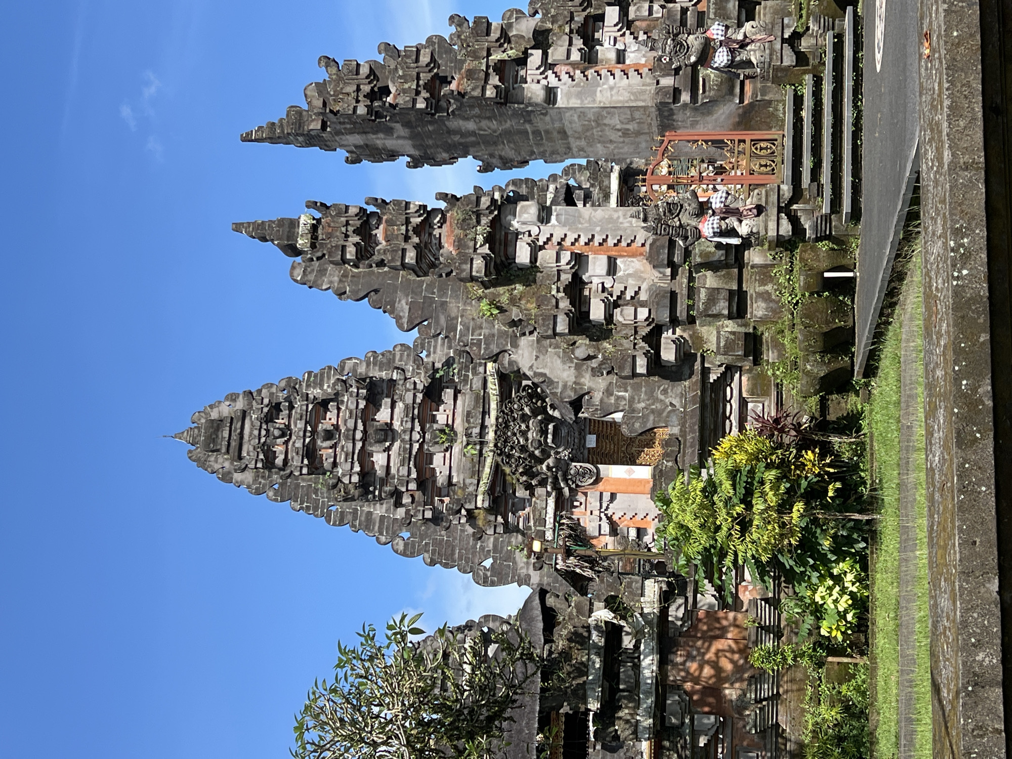 The image shows a Balinese temple in Petulu, Indonesia. Three tall, tiered, stone structures with intricate carvings are visible, set against a clear blue sky. The foreground features lush green vegetation and what appears to be a road with a low concrete barrier. Small figures adorn the temple structures, some wearing checkered cloths. The overall impression is of a well-maintained religious site in a tropical setting.
