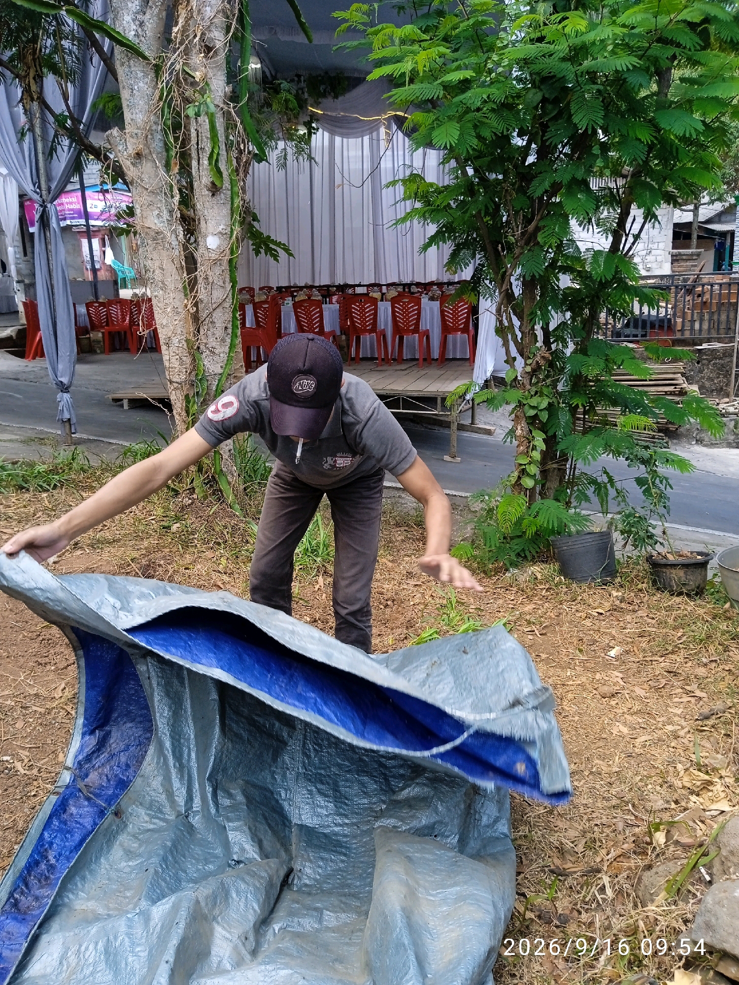 A man in a gray t-shirt, dark pants, and a black baseball cap is crouching on the ground, holding a folded blue and gray tarp. He is partially facing the camera, with his left arm extended and his right hand on the tarp. A cigarette is visible in his mouth. The background shows a pathway with red chairs and tables set up, partially covered by white drapes. There are trees and foliage around the area, and a building with a wooden fence is visible in the distance. The ground is covered with dirt and dry leaves. The timestamp "2026/9/16 09:54" is present in the bottom right corner of the image. The scene appears to be outdoors, possibly at an event or gathering.
