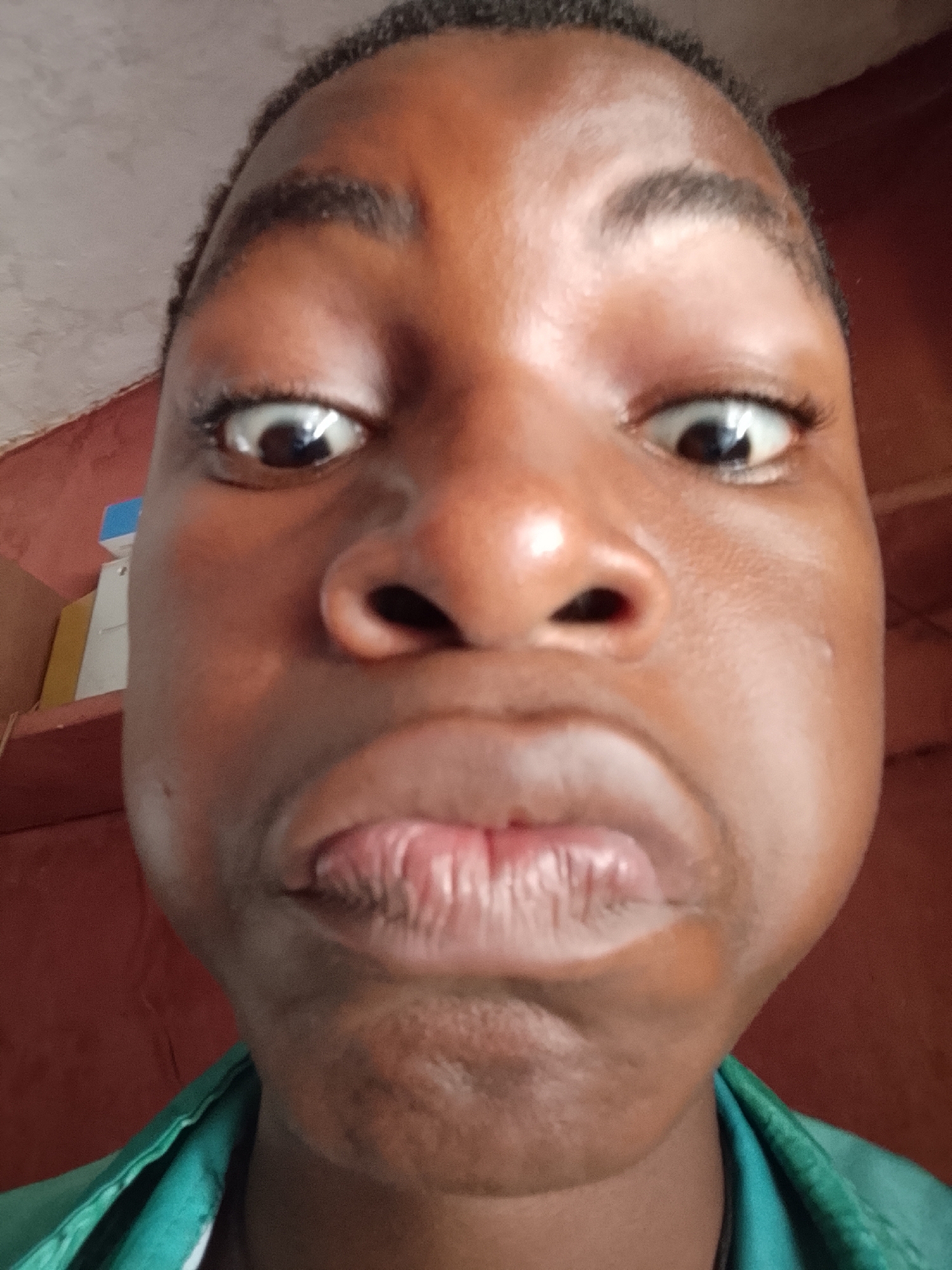 A close-up, front-facing photograph captures the face of a person with dark skin, occupying most of the frame. The perspective is slightly distorted due to the camera's close proximity, suggesting a selfie. The person's eyes are wide open, showing large pupils and a significant amount of white sclera, conveying a look of surprise or an exaggerated expression. Their eyebrows are dark and distinct. The nose is broad, and the mouth is pursed and pushed forward, forming a pronounced pout with visible texture on the lips. The cheeks appear full and rounded.
The person is wearing a garment with a teal or dark green collar visible at the bottom of the frame. The background is simple, featuring a plain wall in shades of reddish-orange or terracotta. To the upper left, a lighter, almost beige or off-white section of a wall or ceiling is visible, meeting the red wall at an angle. On the left side, behind the person's head, there are glimpses of what appears to be a shelf with stacked items, including a blue and white box and a yellow folder or book, indicating an indoor setting. The lighting is bright and even, highlighting the facial features without harsh shadows. The overall emotion conveyed is playful or humorous, as the facial expression seems intentionally exaggerated. No text is visible in the image.