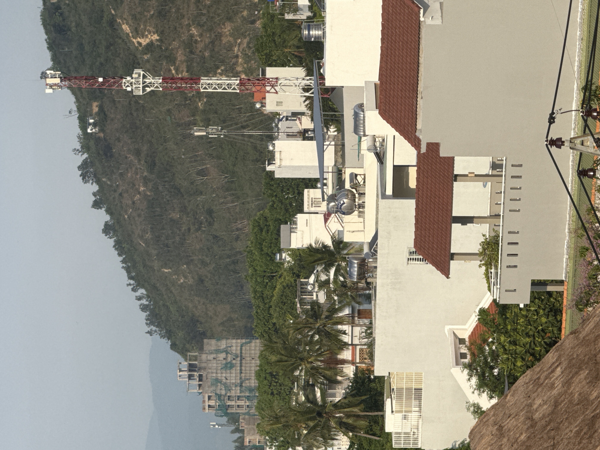 The image shows a telecommunications tower, with red and white lattice structure, situated on a densely wooded hillside. Below the hill, a cluster of white buildings, some with red tiled roofs, are visible. Among these buildings, there are palm trees and construction sites. In the foreground, there are also more buildings with white and grey facades, some featuring tiled roofs. Electrical wires and insulators are also visible in the lower part of the frame. The scene appears to be a residential or mixed-use area in Bac Nha Trang Ward, Vietnam, characterized by a blend of natural landscape and urban development.