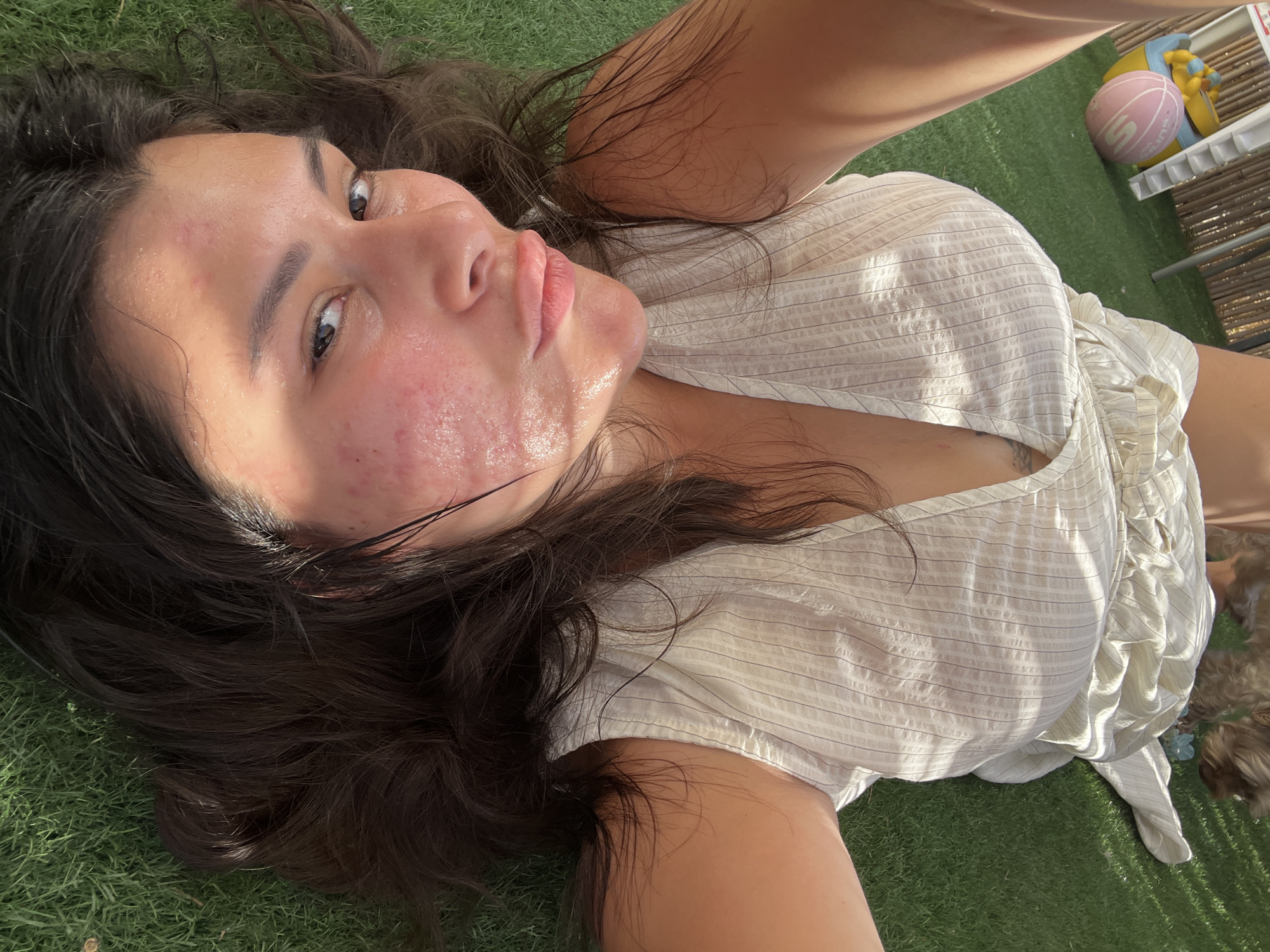 This is a selfie taken from a high angle, looking down at a young woman lying on artificial grass. The woman has long, dark, wavy hair and appears to be looking upwards towards the camera. Her face shows some redness and texture, possibly acne or irritation, and her skin is glistening, suggesting sweat or moisture. She is wearing a light-colored, striped top with a V-neck.
In the background, there are some toys, including a pink ball and a yellow and blue object that appears to be part of a larger toy, possibly a slide. There's also a portion of a bamboo fence or screen. The lighting suggests it is daytime, with sunlight casting shadows. The overall scene appears to be outdoors in a relaxed setting, perhaps a backyard or play area. The woman's expression is neutral. There is no visible text in the image.