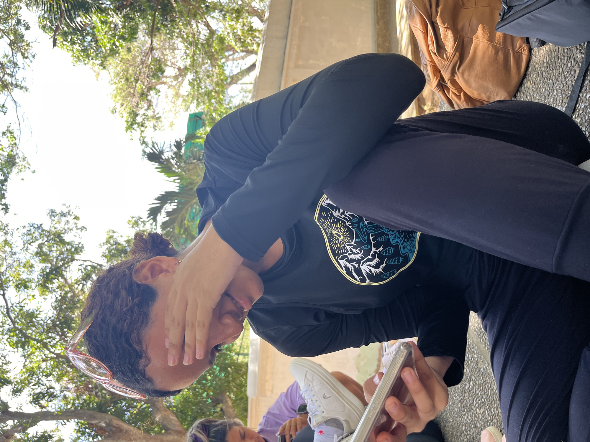 A woman with curly hair pulled into a bun sits outdoors, covering her eyes with her hand and looking down at a smartphone. She wears pink-rimmed glasses perched on her head and a black long-sleeved shirt with a circular design on the chest. She also wears black leggings. A white shoe rests on her lap. Other people are visible in the background, as well as trees overhead. A backpack and a jacket are visible next to her. The ground appears to be paved. The location is El Valle del Espíritu Santo, Venezuela.