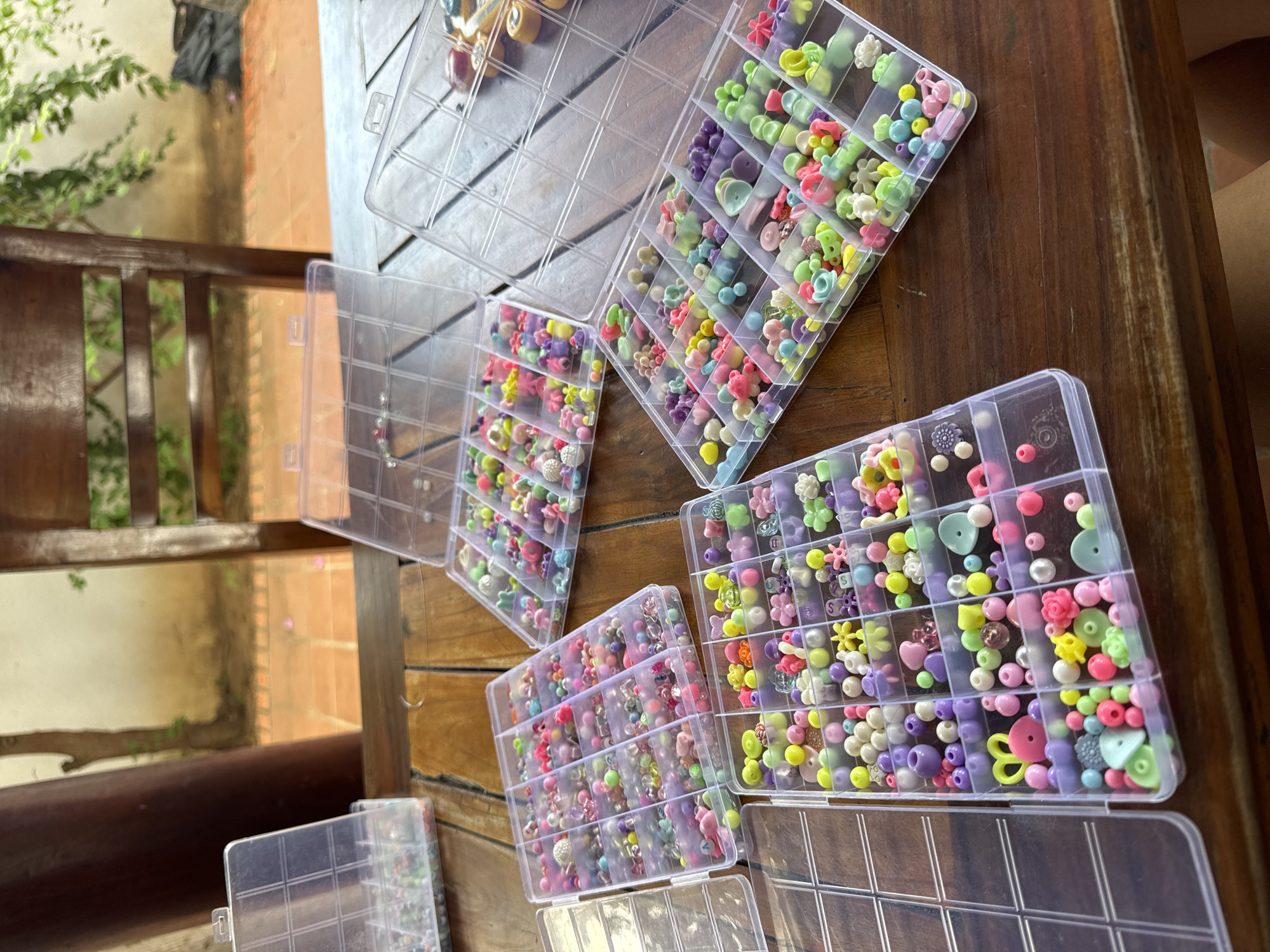 The image displays a close-up, high-angle view of several clear plastic compartmentalized organizers filled with a vibrant assortment of pastel-colored beads and charms. Six organizers are visible on a rustic wooden table, with more implied beyond the frame. Each compartment holds a collection of small decorative items, including round beads, as well as various shapes like flowers, hearts, butterflies, and other intricate forms. The color palette is predominantly soft and cheerful, featuring shades of pink, purple, yellow, green, blue, and white. A single letter 'S' bead is discernible in one of the compartments.
The setting appears to be an outdoor or semi-outdoor space, possibly a patio or balcony. Natural daylight brightly illuminates the scene. In the background to the left, parts of a dark wooden railing or furniture are visible, through which lush green foliage can be seen. Beyond this, a reddish-brown wall or structure and a lighter ground surface suggest an open-air environment. No people are visible, but the arrangement of the beads suggests an ongoing or recently concluded crafting activity, such as jewelry making or other creative projects. The overall mood is one of quiet creativity and organization. The location context of Mũi Né, Vietnam, aligns with an outdoor, sunlit setting.