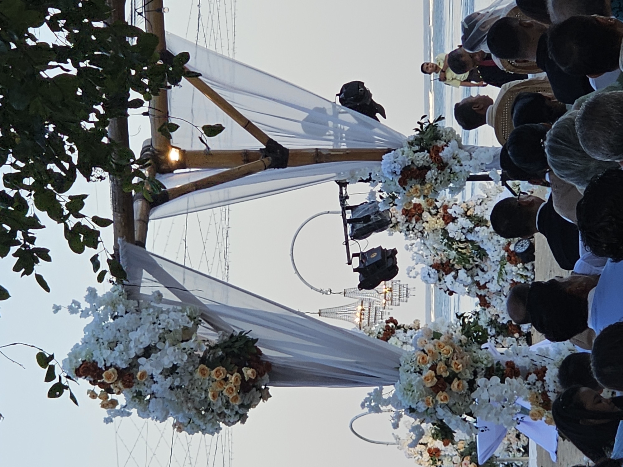 This image captures an outdoor wedding ceremony with a beach setting in Denpasar, Indonesia. The scene is oriented vertically, with the sky at the top and the beach and ocean visible in the background. A bamboo arch, draped with white fabric and adorned with abundant white, cream, and burnt orange floral arrangements, forms the centerpiece of the ceremony setup. Chandeliers and lighting fixtures hang from the arch, suggesting the event might extend into the evening.
Several people are visible, gathered around the arch, though their faces are mostly obscured. One man in a yellow shirt and a man in a white shirt with a patterned collar are more clearly visible, suggesting they might be officiants or key figures in the ceremony. The attire of the guests, primarily dark suits and formal wear, indicates a special occasion. Some individuals are looking towards the front, as if witnessing a vow exchange or a significant moment of the wedding.
The lighting suggests it is late afternoon or early evening, with soft light illuminating the scene. The presence of the ocean in the background clearly places the event on a beach. The overall mood appears celebratory and formal, fitting for a wedding. No visible text is present in the image.