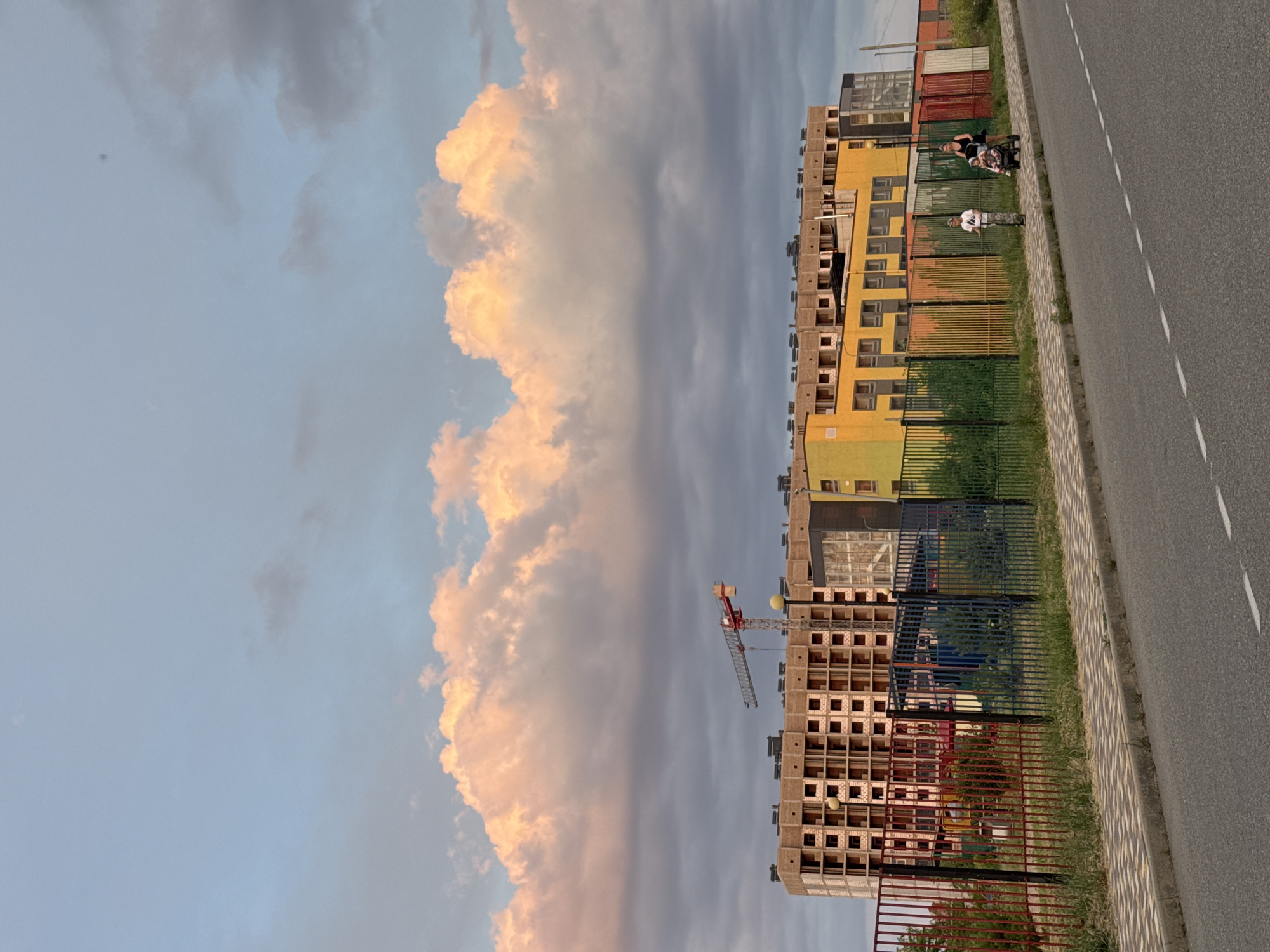 In Mikhaylovsk, Russia, an unfinished multi-story building under construction stands prominently against a sky filled with dramatic, pink-tinged clouds. A red construction crane is visible above the building. In front of the building, a vibrantly colored fence, featuring panels of red, blue, green, yellow, and orange, separates the construction site from a paved sidewalk and road. On the sidewalk, a woman is pushing a stroller with a child, and a child on a scooter is also present. The scene captures a moment of everyday life against the backdrop of urban development.