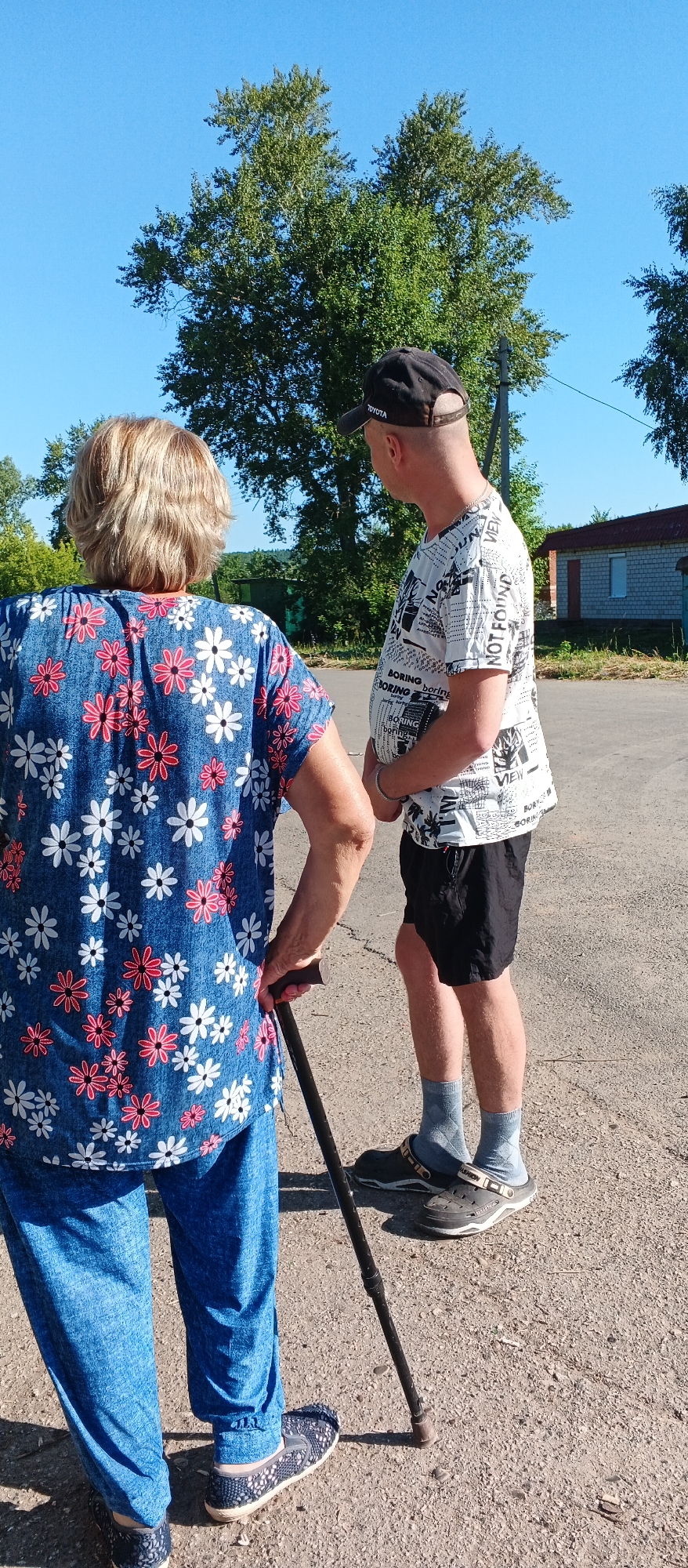 Two individuals are standing on a paved surface outdoors, with a clear blue sky and trees in the background. To the left, a person with blonde, permed hair faces away from the camera, wearing a blue patterned shirt with white and pink flowers and blue pants. They are holding a dark-colored cane in their right hand. To the right, a person wearing a black baseball cap with a "TOYOTA" logo, a white t-shirt with black text and graphics, and black shorts, faces to the left, looking at the other individual. They are wearing light grey socks and dark shoes. In the background, there are trees, a small building with a red roof, and a fence. The environment suggests a rural or semi-rural setting, possibly in or near the village of Подгорное, Russia, given the presence of typical Russian rural architecture and vegetation. The lighting indicates it is daytime with bright sunshine.