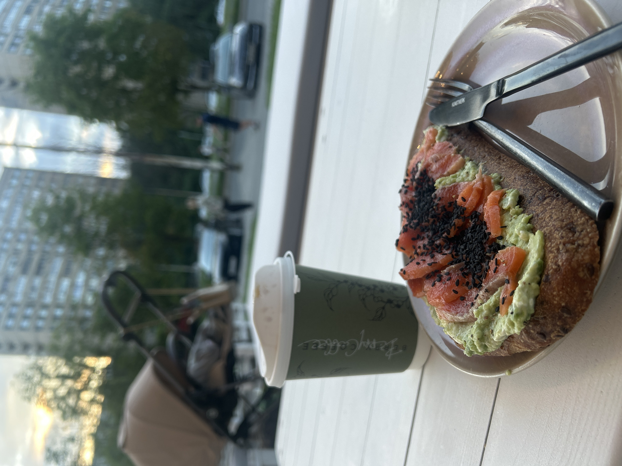 An outdoor cafe scene in Tolyatti, Russia, features a table with a slice of avocado toast topped with salmon and black sesame seeds, a green paper coffee cup with a white lid, and cutlery on a light brown plate. In the blurred background, a stroller, trees, apartment buildings, and parked cars are visible, suggesting a residential area. The lighting indicates it is either early morning or late afternoon.