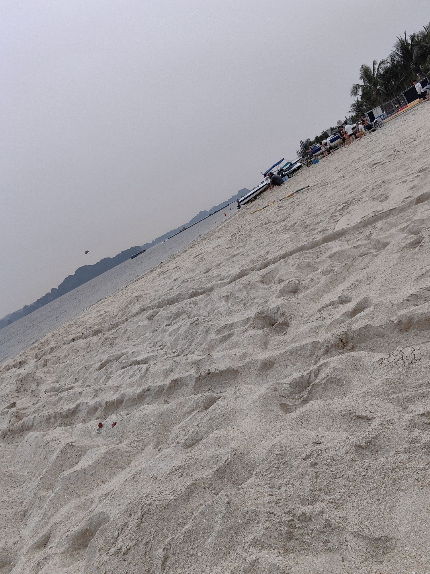 The image shows a beach scene in Bai Chay Ward, Vietnam. The foreground is dominated by a wide expanse of sandy beach. In the distance, the ocean meets a hazy sky. Several people and boats are visible on the beach. Palm trees line the edge of the beach. There is also a ship.