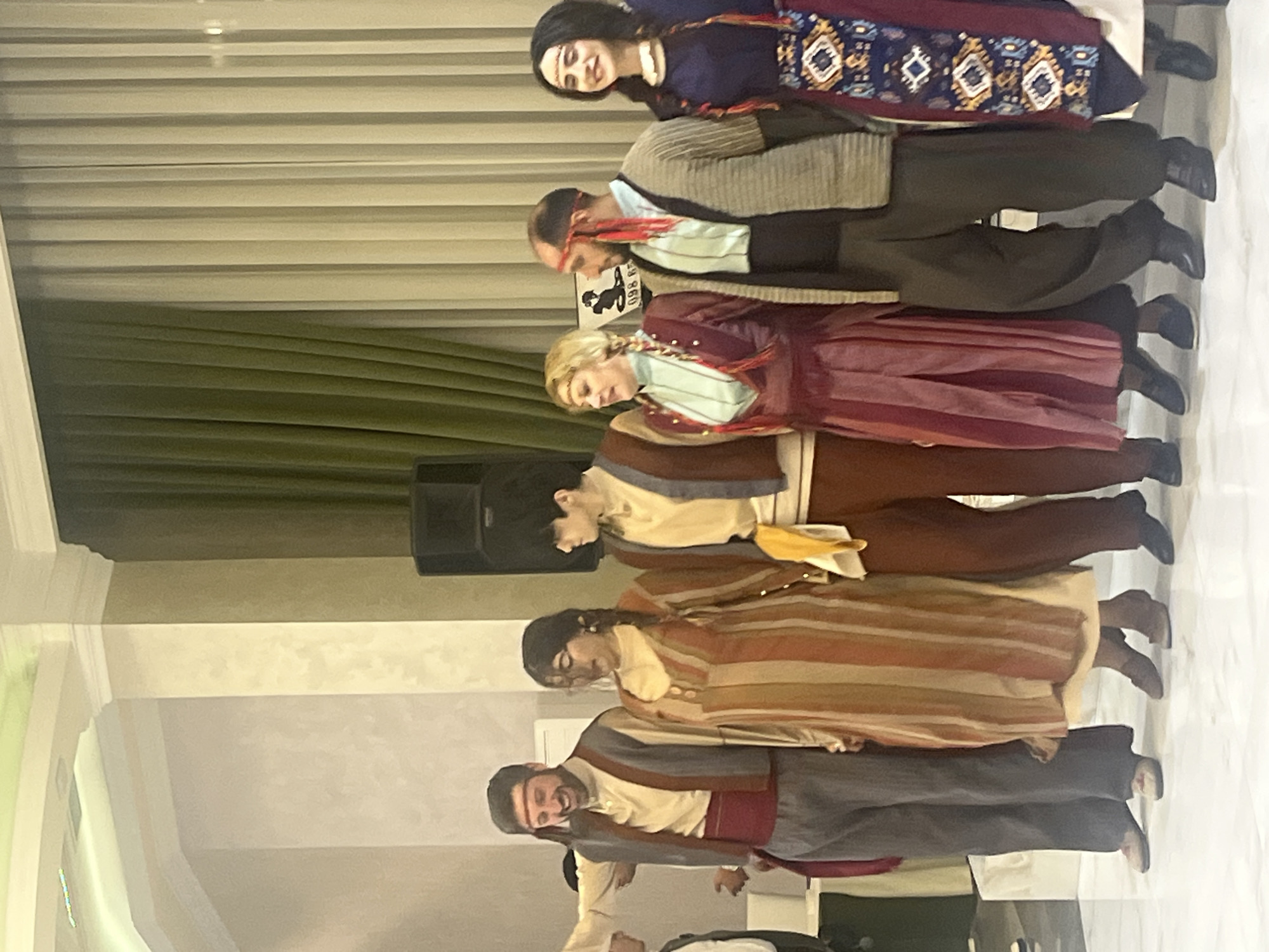 In Areni, Armenia, an indoor scene depicts seven individuals, four males and three females, standing in a horizontal line. All subjects are adults and are attired in distinct traditional or folk costumes.
From left to right:
1. A male, partially visible, wearing a light-colored long-sleeved shirt, a dark vest, and dark trousers.
2. A smiling male with a beard, wearing a red headband, a light-colored long-sleeved shirt, a greyish-blue vest with brown trim, a red waist sash, dark grey trousers, and light-colored shoes. His right arm is raised.
3. A female with braided dark hair and glasses, clad in a long, vertically striped dress in hues of brown, orange, and cream. She wears brown low-heeled shoes.
4. A male with dark hair, wearing a light-colored long-sleeved shirt, a dark reddish-brown vest with light trim, dark reddish-brown trousers, and carrying a yellow cloth on his left side. Dark shoes are visible.
5. A female with blonde braided hair adorned with red accents, wearing a light blue long-sleeved top beneath a dark red long dress. She wears dark low-heeled shoes.
6. A male with short hair or bald, a beard, and a red headband featuring red tassels. He is dressed in a light blue long-sleeved shirt under a dark top or cardigan, dark trousers, and dark shoes.
7. A smiling female with braided dark hair also adorned with red accents, wearing a dark blue long dress with white trim at the neckline and a skirt portion patterned with geometric designs in white, orange, blue, and black. Dark shoes are visible.
The background consists of light-colored, possibly textured walls with white crown molding near the ceiling. Green and light beige vertical-pleated curtains are visible on the right side, behind which a large black speaker is positioned. A small, partially visible sign with the text "098 RE..." is located behind the fifth and sixth individuals. The floor is light-colored and reflective, suggesting a polished surface. The lighting is bright and even, consistent with an indoor event space.