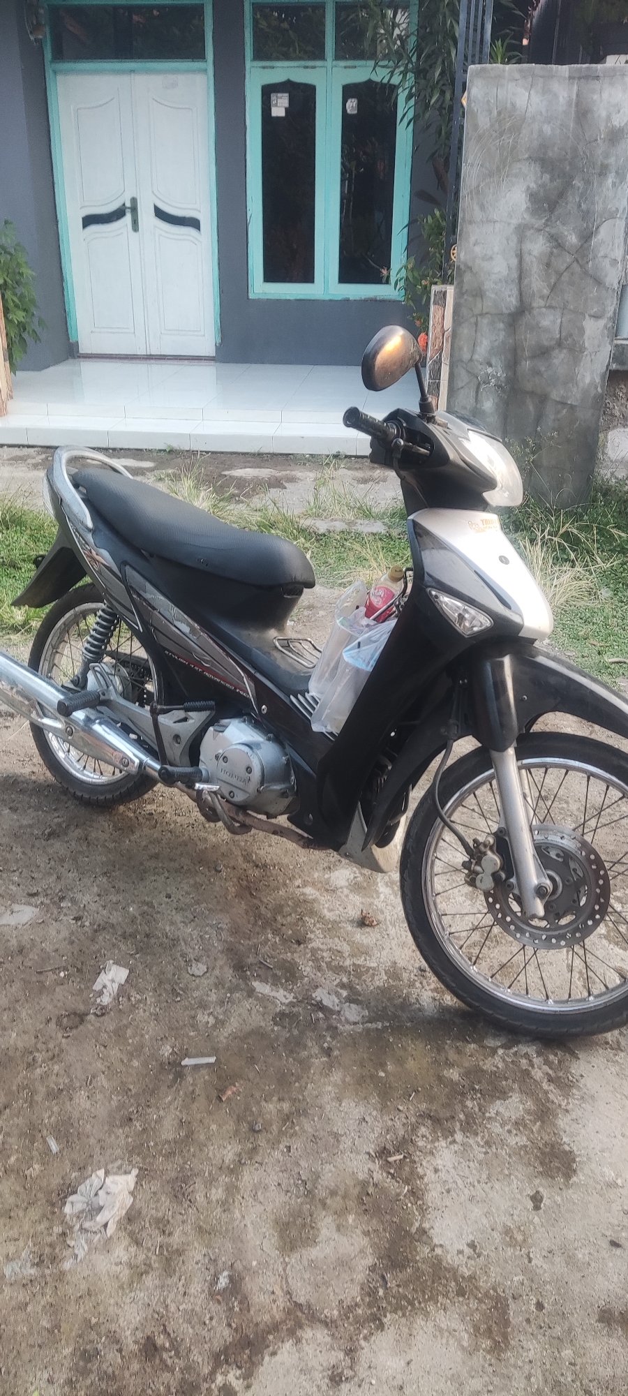 A black and silver motorcycle is parked on a dirt and gravel surface in front of a building. The motorcycle has a black seat, silver trim, and spoked wheels. A clear plastic bag with some items inside is attached to the handlebars. In the background, there is a white double door and a window with turquoise-colored trim. To the right of the motorcycle, a rough, grey concrete wall is visible. The ground is uneven and appears to be a mix of dirt, small stones, and some scattered debris, including pieces of paper. The scene is outdoors, likely in a residential or yard area in Bendungan, Indonesia.