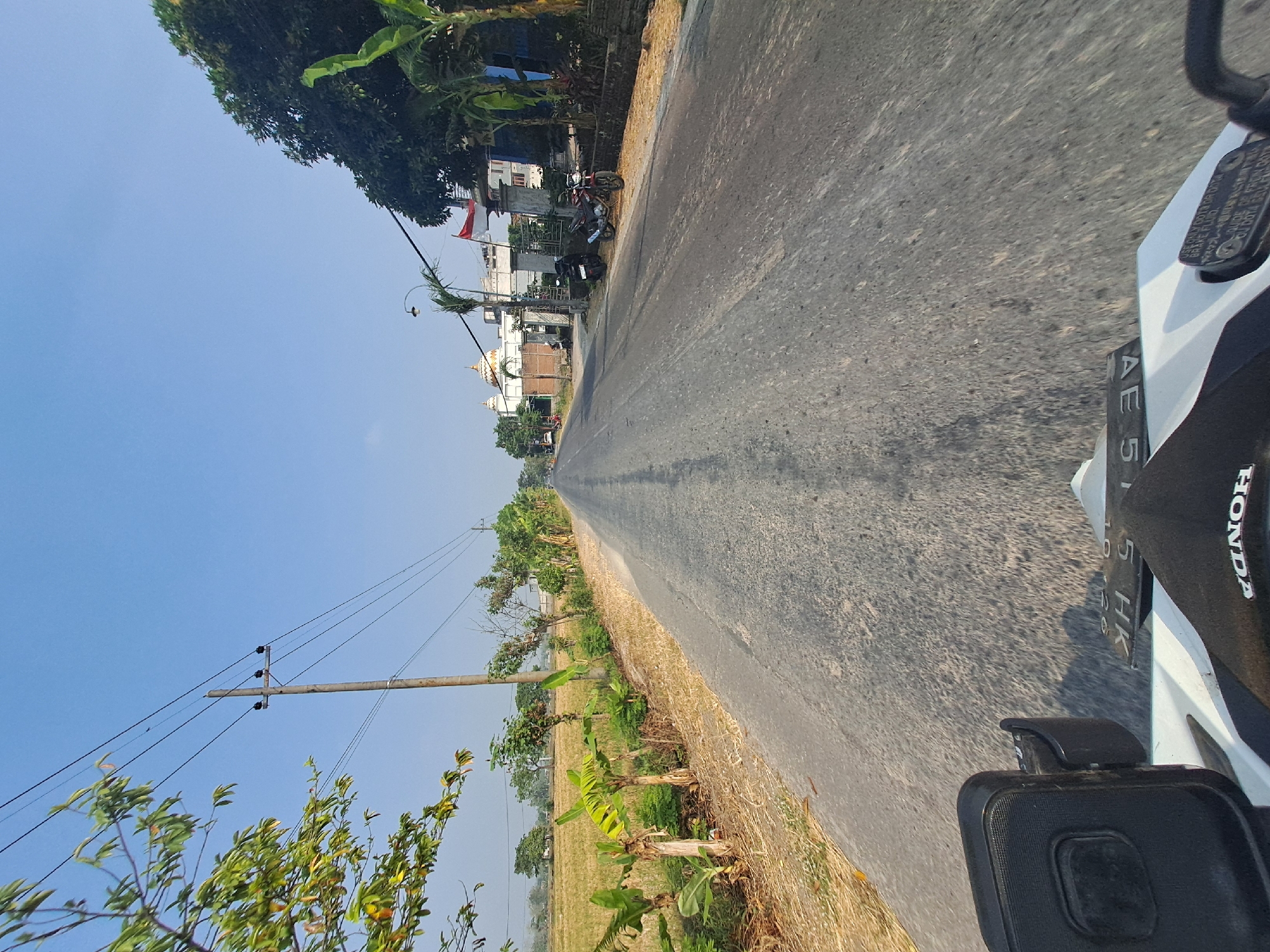 The image captures a view from a motorcycle on a road in Singgahan, Indonesia. The road stretches into the distance, flanked by agricultural fields on the left and trees and buildings on the right. A utility pole with electrical wires stands prominently on the left side of the road. In the distance on the right, a white building with domes is visible, possibly a place of worship. Two motorcycles are parked on the side of the road near the building. The sky is clear and blue, indicating a sunny day. The motorcycle's front portion, including a black seat and part of the white bodywork with a license plate, is in the foreground.
