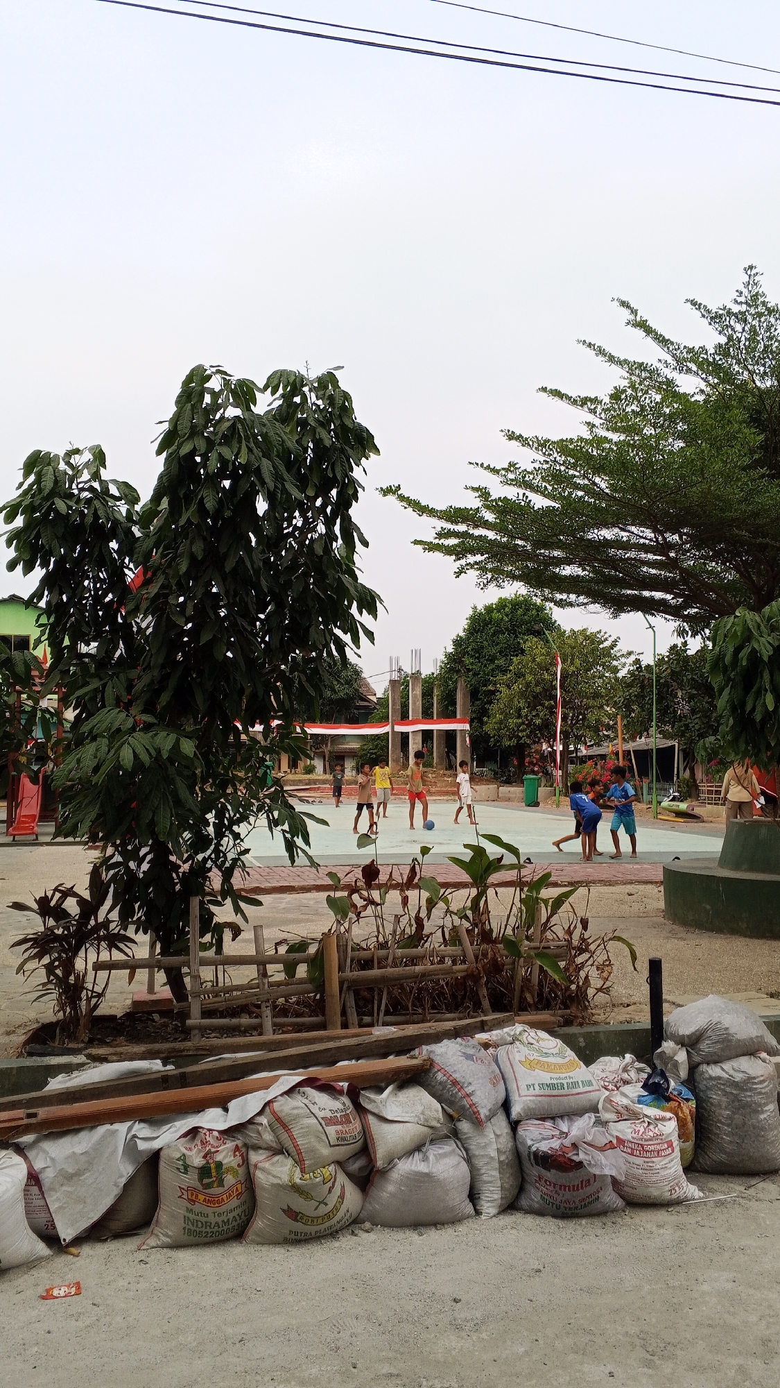 In an outdoor setting, possibly a public space or schoolyard in Bekasi, Indonesia, a group of young boys are playing soccer on a paved court. The court is surrounded by trees and buildings, and a red and white Indonesian flag is visible in the background. In the foreground, several large bags, likely containing construction materials or waste, are piled up on the ground. The sky is overcast, suggesting a cloudy day. The overall scene depicts a casual, everyday activity taking place amidst signs of ongoing development or renovation.