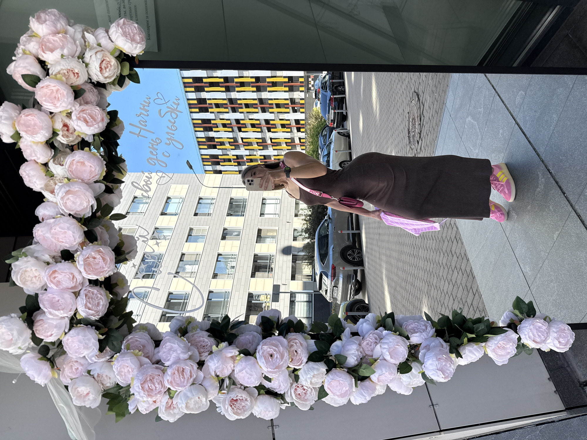 A young woman stands in front of a building with a colorful, geometric facade, reflected in a large mirror adorned with a floral arch made of pink and white peonies. The woman is wearing a long, dark brown dress and bright pink sneakers. She is holding a pink bag and a smartphone, taking a selfie. The mirror also shows a clear blue sky and some text written in Russian, which translates to "Start your day with a smile." The location appears to be Saint Petersburg, Russia, based on the architectural style and the Cyrillic text.