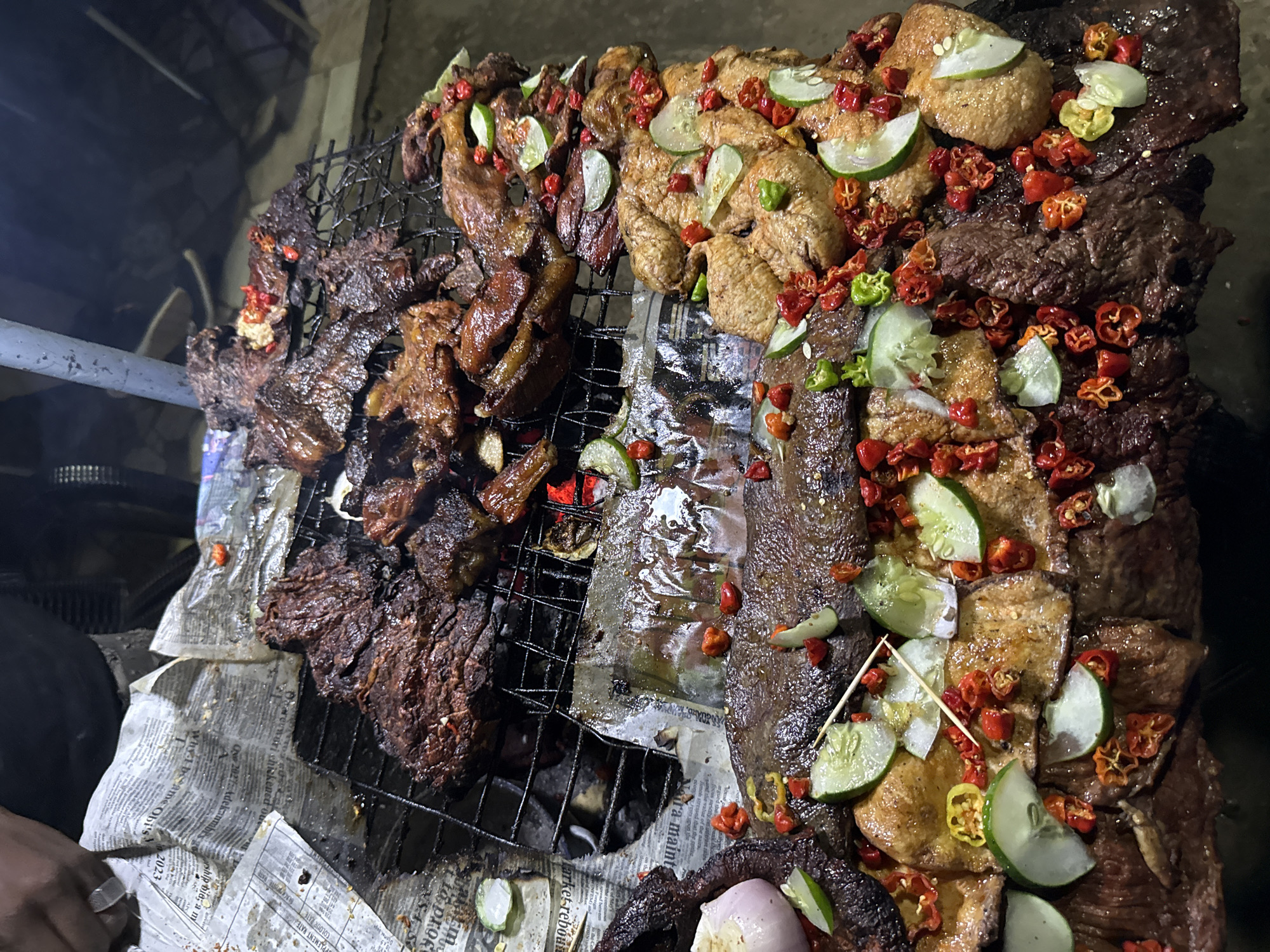 The image shows a close-up, high-angle view of a variety of grilled meats and fish on a metal grate over coals. The food is seasoned with chopped chili peppers and garnished with slices of cucumber and what appears to be a piece of onion. The grate is placed on top of a pile of newspapers, which are tattered and have some smudges on them. In the upper left corner, a person's arm and hand are visible, wearing a ring on one finger, suggesting they are involved in the preparation or serving of the food. The lighting is dim, indicating it may be evening or nighttime. The overall scene suggests an outdoor food stall or informal cooking setup, likely in a market or street setting. While the image does not explicitly show the city of Abuja, Nigeria, grilled street food is a common sight in many Nigerian cities, including Abuja, particularly in areas like Wuse Market or Garki Market, where vendors often sell these types of delicacies.