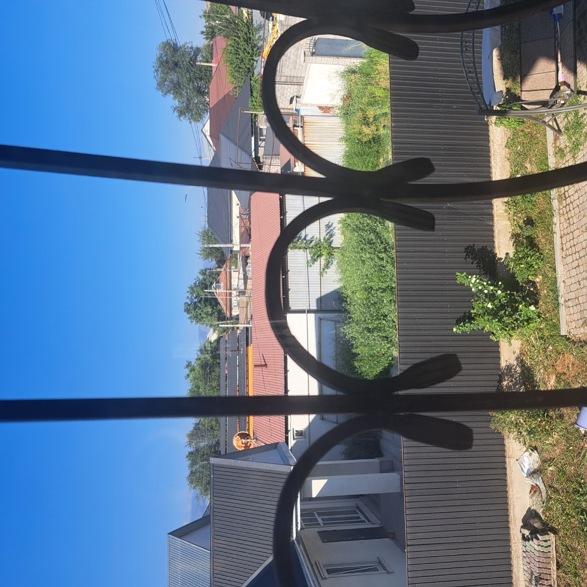 The image shows a view through a decorative metal fence of a residential area under a clear blue sky. In the foreground, the dark, wrought-iron fence's curved decorative elements are prominent. Beyond the fence, several houses with various roof colors, including red and gray metal, are visible. Green trees and foliage are interspersed among the buildings. A dark, corrugated metal fence runs horizontally across the mid-ground. In the lower foreground, a patch of grass and some paving stones are visible. A metal swing set is partially visible on the right side of the image. The location is identified as Іргелі ауылдық округі, Kazakhstan.