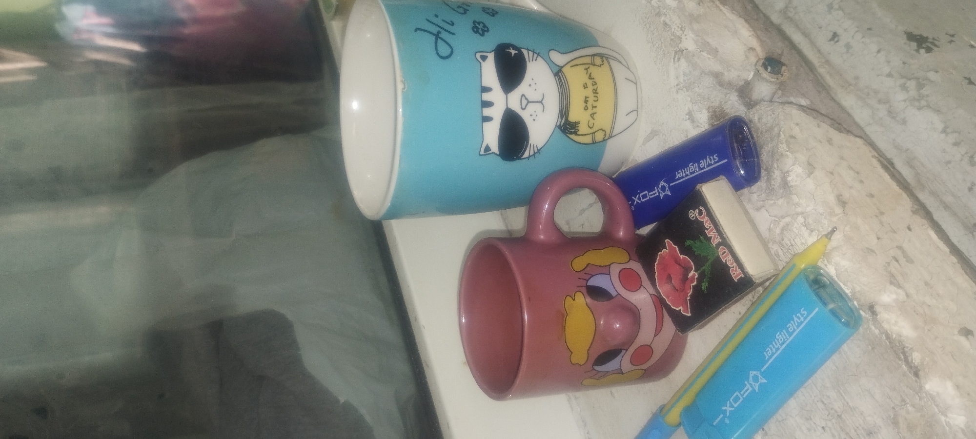 This image displays a collection of objects arranged on a textured, light-colored surface, possibly a windowsill or a rough concrete ledge. Two ceramic mugs are the central subjects. The first, a light blue mug, features a graphic of a cat wearing sunglasses and a yellow shirt with the text "DAY TO CATUREDAY" printed on it. The second mug is a pinkish-red color and has a cartoon face printed on it, with large blue eyes, red cheeks, and yellow hair.
Several writing implements and small items are scattered around the mugs. A blue cylindrical object with the text "style lighter" and "FOX" is visible, as is a yellow pen. A small rectangular item, possibly an eraser or a matchbox, has an image of a red flower and the text "Red Mac" on it. The surface appears weathered and has cracks and dark patches, suggesting it might be outdoors or in an unkempt area. The lighting is dim, casting subtle shadows and giving the scene a slightly grainy quality. There are reflections in the background, indicating a possible window or glass surface nearby. The overall impression is of everyday items casually placed together. The presence of "Zaporizhzhia, Ukraine" in the location context hints at a possible geographical setting for this arrangement of objects.