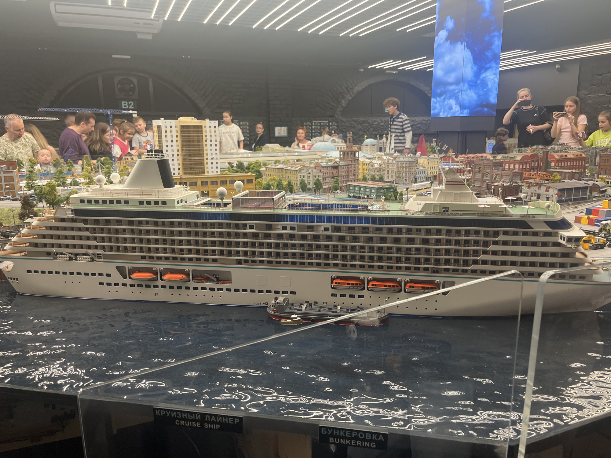 The image shows a detailed diorama of a large cruise ship docked in a miniature city. The cruise ship is white with multiple decks and numerous windows. Orange lifeboats are visible along its side. In the water beside the ship, there is a smaller tugboat and a barge. The diorama also includes various miniature buildings, trees, and street details, resembling a city. In the background, several people, including adults and children, are observing the diorama. Some are looking at their phones, while others are pointing or talking. The setting appears to be an indoor exhibition or museum, given the lighting and the display structure. A sign in the foreground reads "КРУИЗНЫЙ ЛАЙНЕР CRUISE SHIP" and "БУНКЕРОВКА BUNKERING." The diorama is located at the Museum of the Russian Navy in Kronstadt, a city within the federal city of Saint Petersburg, Russia.