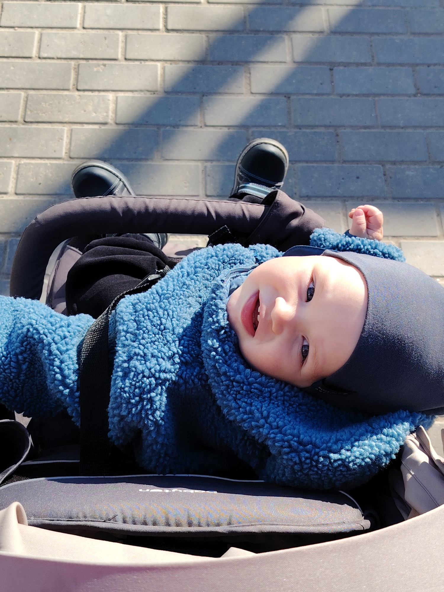 A baby lies in a stroller on a brick sidewalk. The baby is wearing a blue fleece jacket and a dark blue beanie. The baby is smiling and looking up at the camera. The stroller has a black handlebar and a grey seat. The sidewalk is made of grey bricks and there are shadows cast on the bricks. The location cannot be confirmed as Chaikovsky, Russia, from this image.