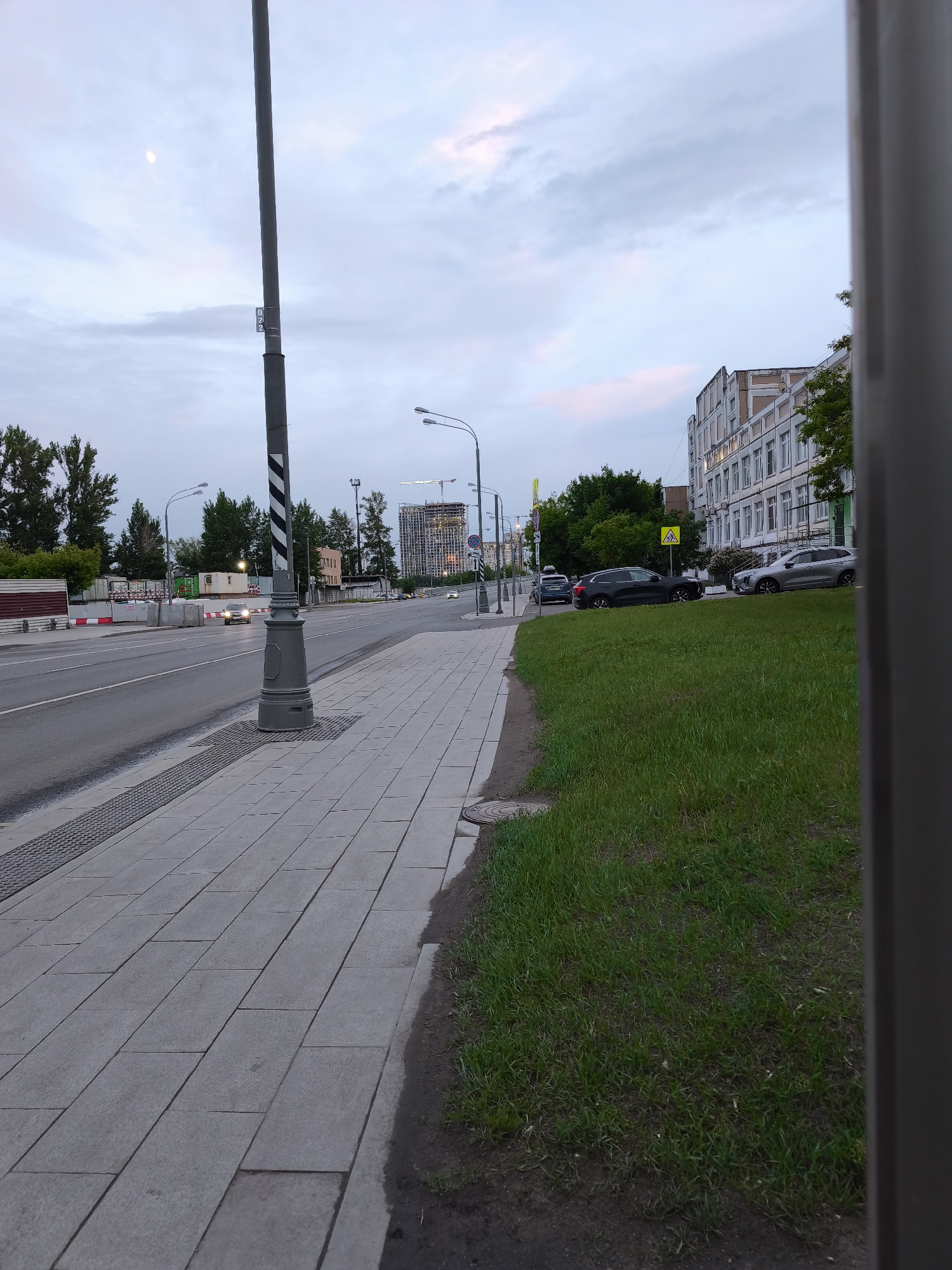 The image shows a street scene in Moscow, Russia at dusk. The foreground features a gray-tiled sidewalk with a gray light pole on the left. To the right of the sidewalk is a grassy area. In the background, a street with cars can be seen. Buildings, including a white, multi-story building, are visible on the right. A construction site with a crane is also visible in the distance. The sky is cloudy with hints of pink and blue.
