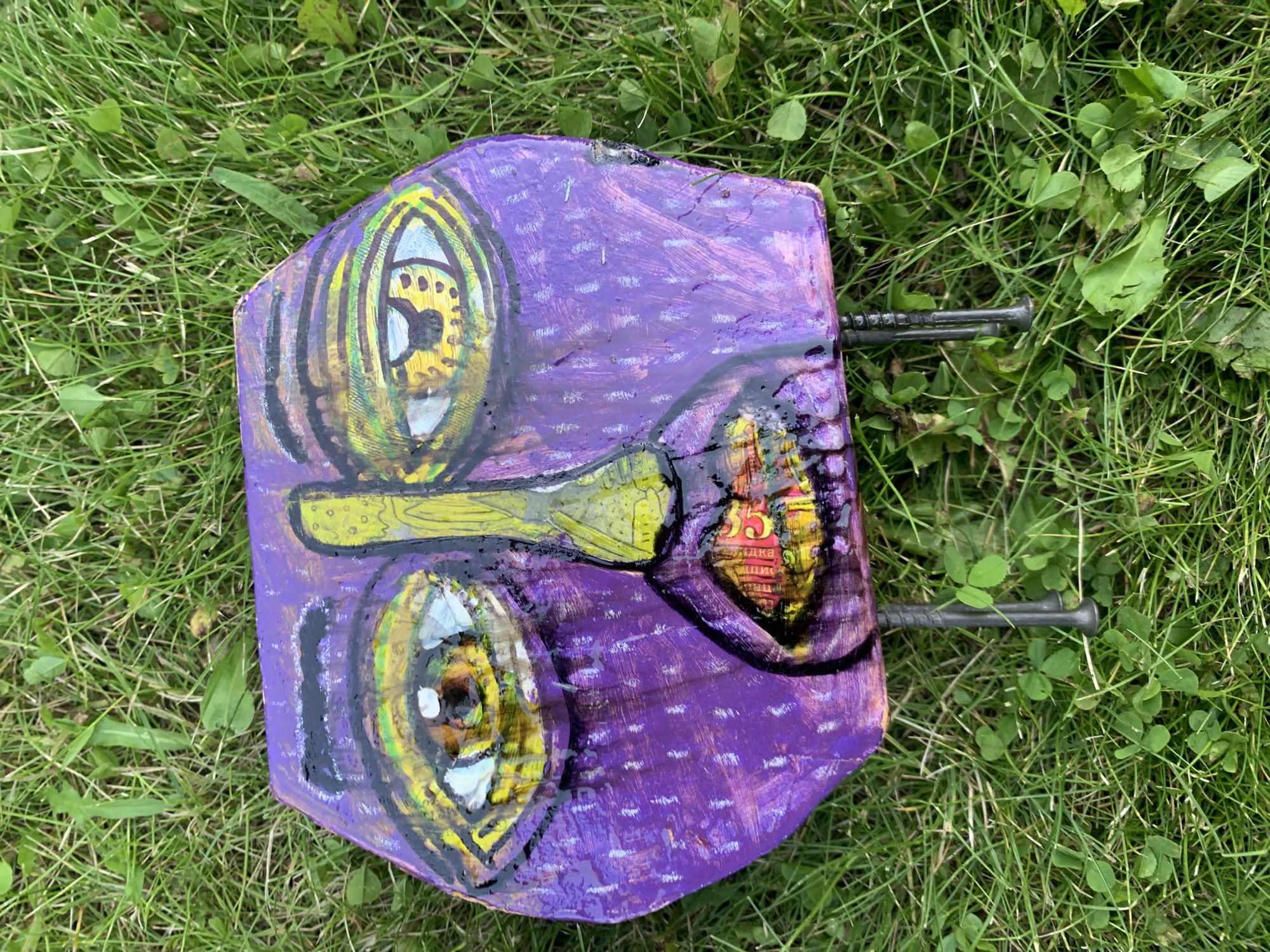 This is a close-up, overhead shot of a folk art sculpture, resembling a stylized face, resting on a patch of green grass. The sculpture is hexagonal and painted primarily in vibrant purple. It features large, expressive eyes with yellow irises and black pupils, outlined in black. A prominent yellow nose is painted down the center of the face, and a wide mouth with red and yellow lips is depicted. The sculpture has two thin, dark metal legs that support it. The background is entirely composed of green grass with some small, lighter green leaves scattered throughout. The lighting suggests it is daytime, with shadows present but not harsh. The location is not specified in the image.