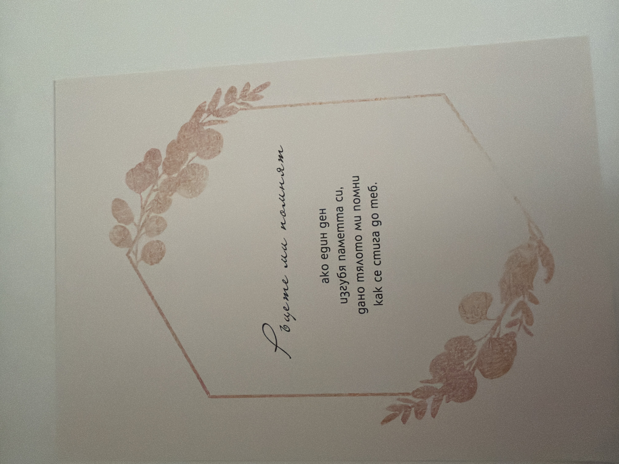 The image shows a printed card with decorative floral elements and text. The text appears to be in Bulgarian. The card is light-colored and has a faint reddish-pink floral design framing the text. The design consists of abstract leaf-like shapes. The text is divided into two parts: a handwritten-style font at the top and a more standard font below. The bottom text reads: "ako edun den izgugya pametma cu, daño tyaloto mi pomnu kak se stuga do teb." The top text is in a cursive font and is difficult to decipher fully but appears to be related to memory. The image is taken from a slightly elevated angle, showing the card resting on a plain surface, likely a table or desk. There are no people or external environmental elements visible. The location of this card in Pazardzhik, Bulgaria is not discernible from the image itself.