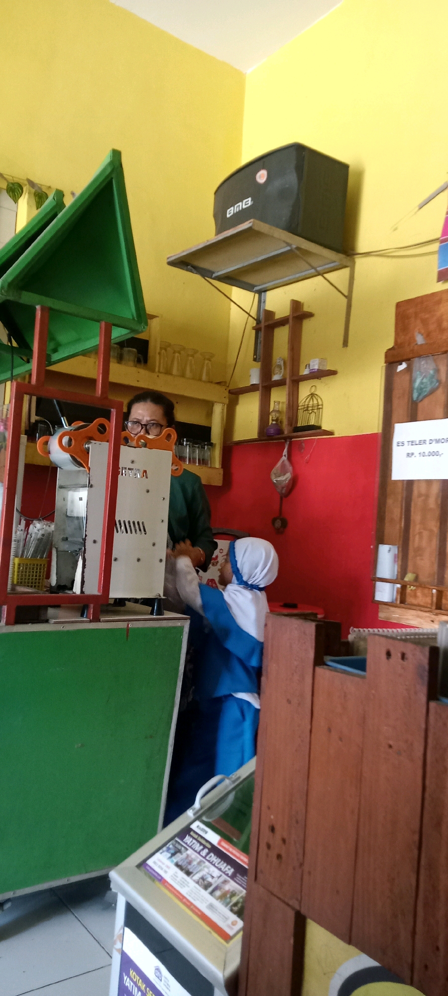 This is a medium-shot, eye-level, indoor photograph taken in the city of Pondok, Indonesia. The scene appears to be a small shop or food stall with a bright yellow wall in the background. In the foreground, to the left, is a green structure with a red frame. A person wearing glasses and a dark green shirt stands behind a machine with a white panel. To the right, a person wearing a blue and white uniform, including a headscarf, interacts with the machine. A speaker is mounted on the wall above a shelf. Shelves with glasses and decorative items are visible on the right side of the image, against a red wall. A sign with text in Indonesian and numbers is posted on a wooden surface on the far right. The overall impression is of a busy commercial space.