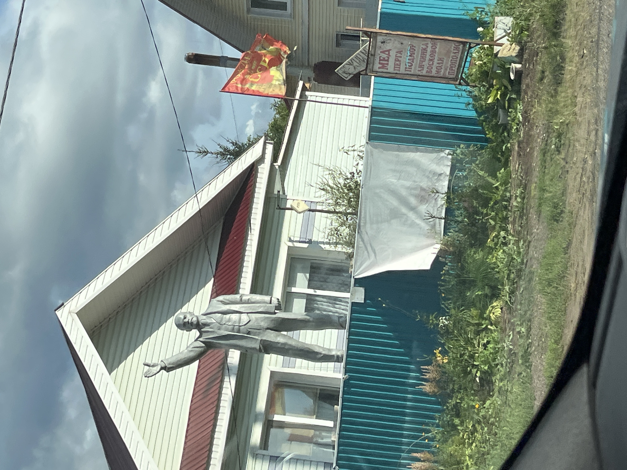 A life-size, gray statue of Vladimir Lenin stands with his right arm raised in front of a light-colored, two-story house with a red-shingled roof. The statue is positioned on top of a teal-colored corrugated metal fence. A red and yellow flag, bearing Cyrillic script, is attached to a pole to the right of the statue, also in front of the house. A sign with Cyrillic text is visible near the flag. The environment is rural, with green foliage and some scattered wildflowers in front of the fence. The sky is overcast with clouds. The image appears to have been taken from a vehicle, suggested by the black foreground element which resembles a car's dashboard. The location is identified as Kryukovo, Russia.