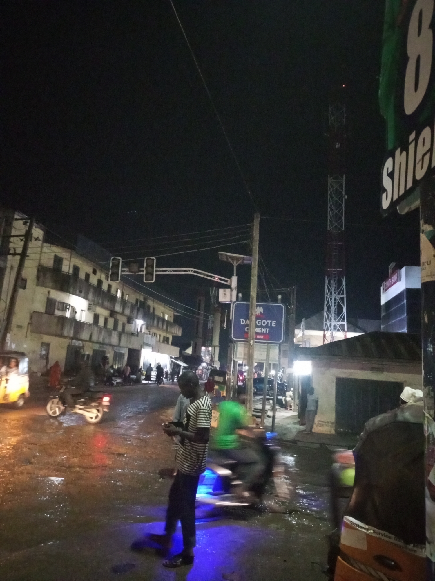 This night-time photograph captures a bustling street scene in Maiburiji, Nigeria, characterized by artificial lighting against a dark sky. The ground is visibly wet and reflective, suggesting recent rainfall or standing water on the asphalt.
Numerous people are present, either standing on the roadside or walking along the sidewalks. Several motorcycles (okadas) are visible; one on the right side of the frame shows significant motion blur, indicating it is moving. A yellow auto-rickshaw (keke napep) is partially seen on the far left.
The street is flanked by buildings. On the left, a multi-story structure with multiple balconies and illuminated windows stands prominently. On the right, a tall, red and white communication tower dominates the skyline, featuring a small red light at its top. Further along, a modern building with "ZENITH BANK" clearly visible on its facade is present.
Traffic lights are suspended over an intersection, and various commercial signs add to the urban environment. A blue sign prominently displays "DAGOTE CEMENT" (likely DANGOTE CEMENT). On the far right, a green sign shows a large number "8" and the partial word "Shield." The scene depicts a typical busy street evening with active transportation and people moving about.