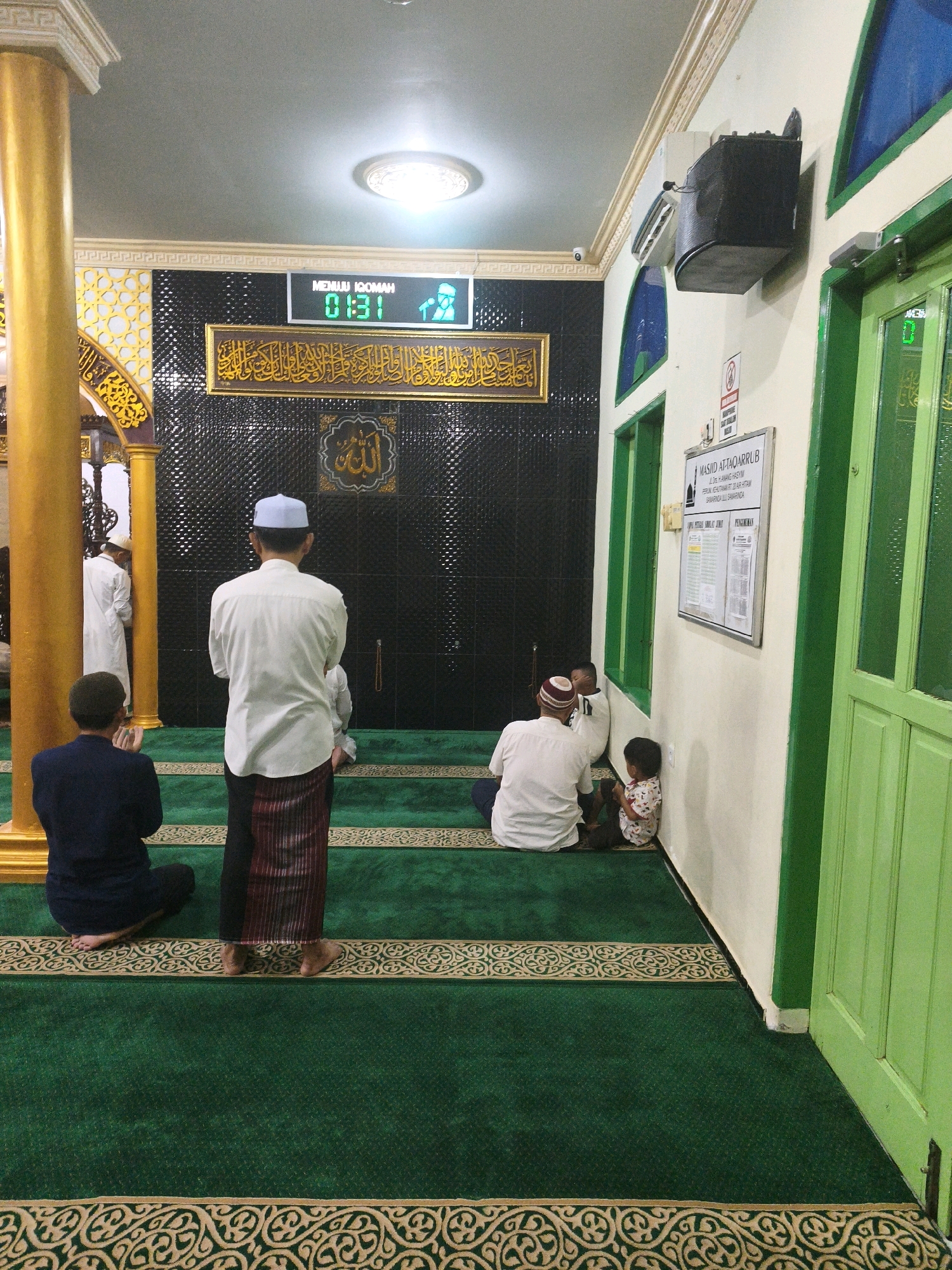 The image shows the interior of a mosque in Samarinda, Indonesia. The mosque's flooring is carpeted in green with ornate gold patterns. Several individuals are present, including adult men and at least one child. In the foreground, a man in a dark blue shirt and trousers is kneeling on the carpet, facing away from the camera. To his right, another man in a white shirt and a sarong is standing, also facing away. Behind them, closer to the back wall, other individuals are seated or kneeling. A digital display above the back wall shows "MENUNU IQOMAH" and the time "01:31", along with an icon of a person. The back wall is black and textured, with a golden frame containing Arabic calligraphy. To the right, a door painted green is visible, along with a white wall that has a notice board with some papers attached. The scene suggests a prayer or congregational gathering is about to take place or is in progress.