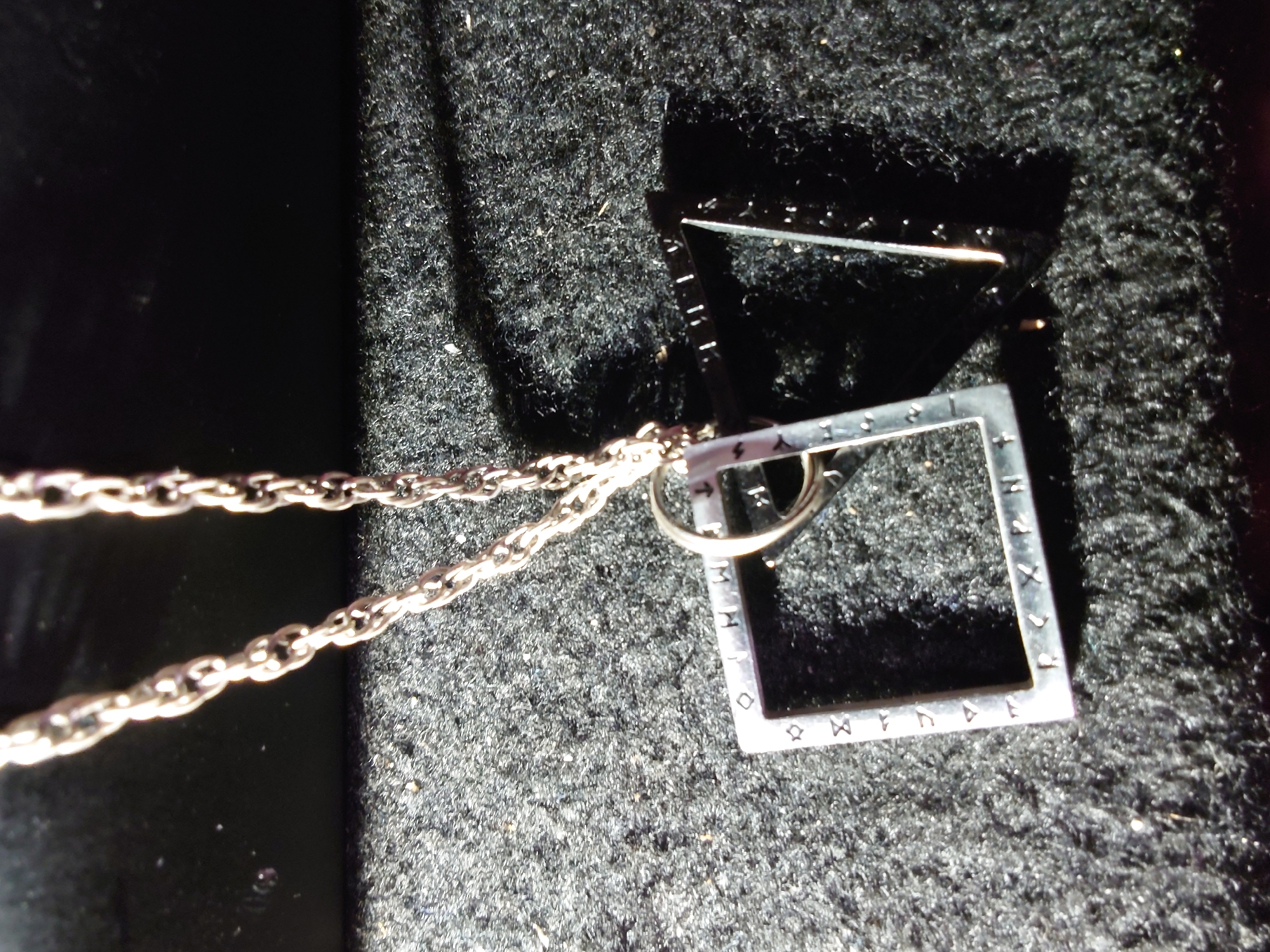 A silver chain with a square pendant is displayed on a dark, textured surface, possibly fabric. The pendant has a hollow interior and appears to have symbols or markings etched along its edges. A second, triangular frame is positioned partially behind and to the right of the square pendant, casting a shadow. The lighting in the image creates strong contrasts and highlights, suggesting an artificial light source. The scene is static and shows no signs of human presence or activity. The location in Sokolohirsk, Ukraine, is not visually indicated in the image.