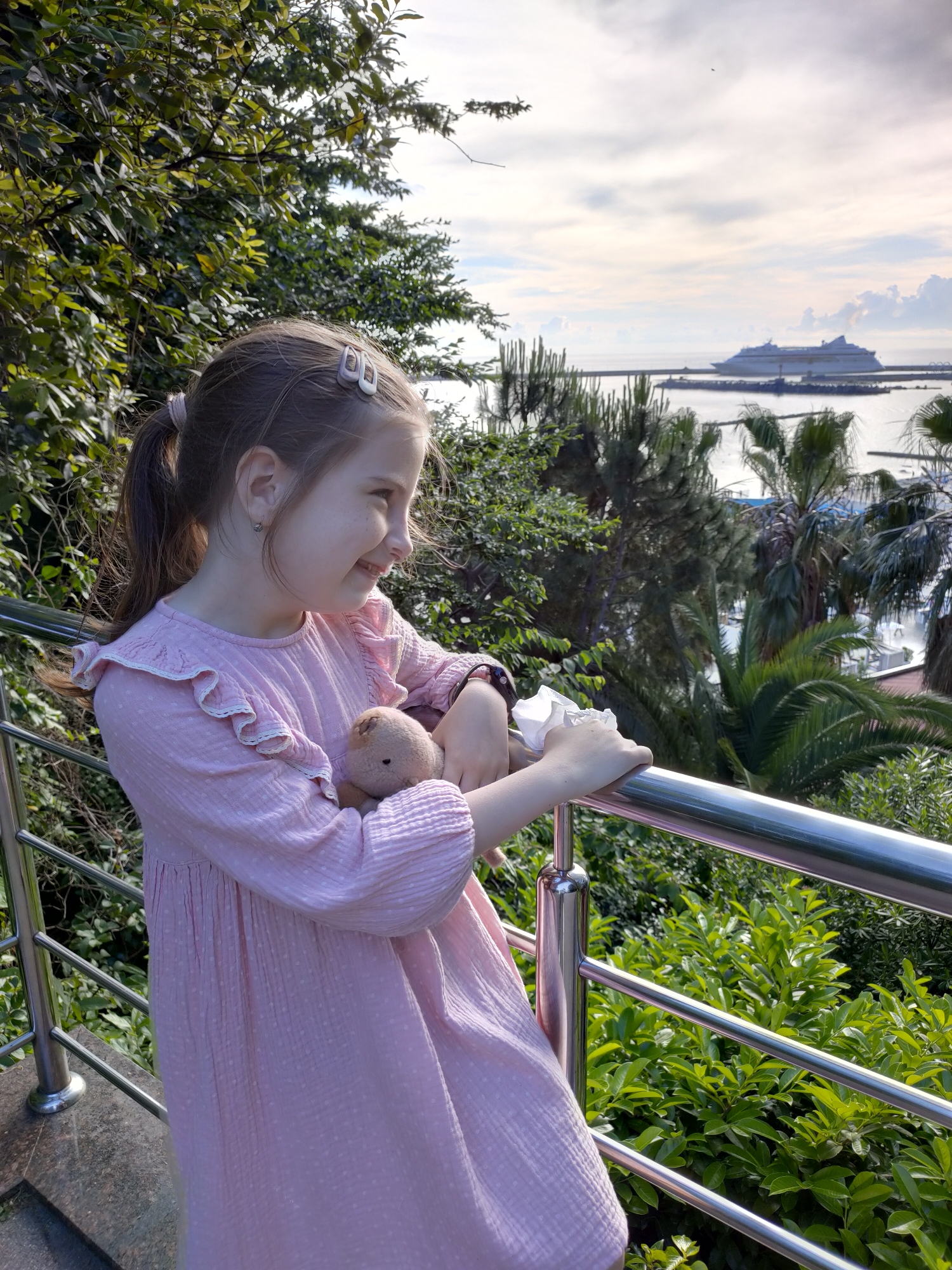 A young girl with brown hair pulled into a ponytail and two small hair clips is looking to the right with a smile. She is wearing a pink dress with ruffled sleeves and is holding a small stuffed animal and a white tissue. She is standing by a metal railing. In the background, there is a body of water with a large cruise ship and palm trees. The scene appears to be in Sochi, Russia, a resort city on the Black Sea coast, known for its warm climate and numerous cruise ship terminals. The image is taken outdoors during daylight.