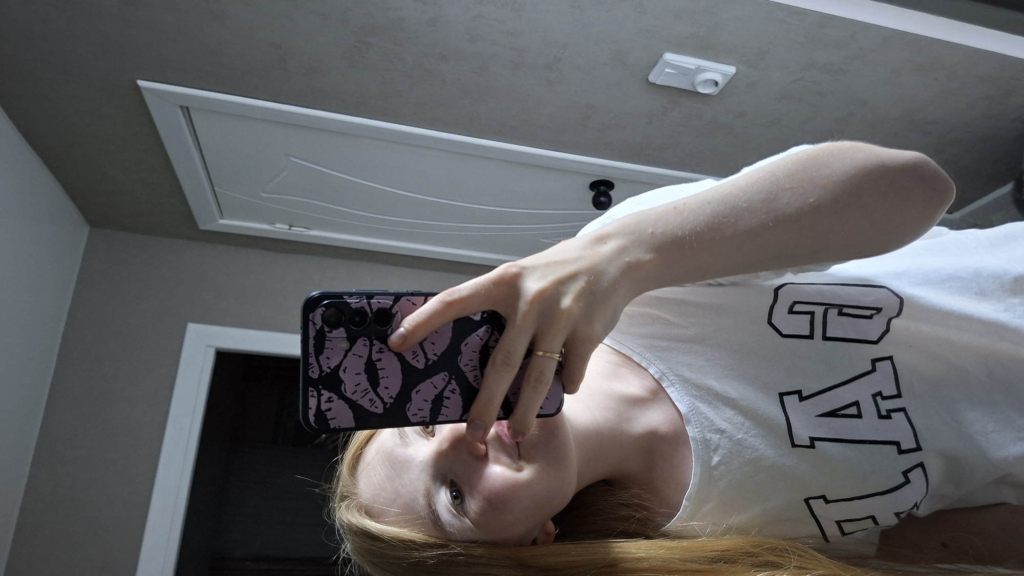 A young woman with blonde hair is taking a selfie in a mirror. She is wearing a white t-shirt with the word "GAP" printed on it in large, black and white letters. Her phone, which has a dark case with a pattern of pink lips, is held up to take the picture. The background appears to be an indoor setting, possibly a bathroom, with a grey wall, a white door, and a doorway to another room visible. The lighting suggests it is daytime. The location is identified as the city of Chaikovsky, Russia, based on context.