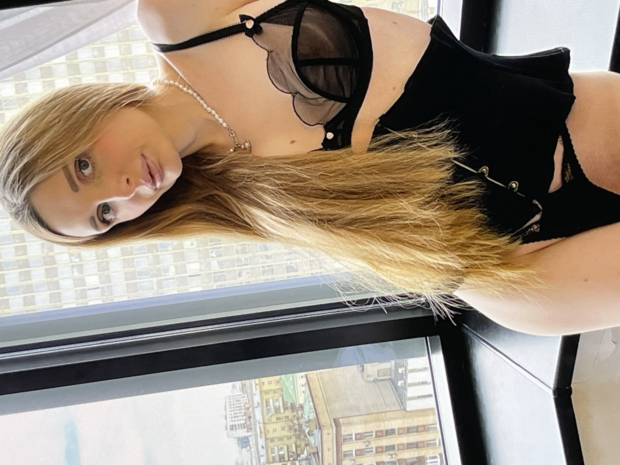 A woman with long blonde hair is posing by a window in what appears to be a high-rise apartment in an urban setting. She is wearing black lingerie, including a bra with sheer panels and a corset. Outside the window, buildings of various heights and architectural styles are visible, suggesting a city environment. The lighting appears natural, coming from the window, and casts soft shadows. The overall impression is one of a candid or posed photograph taken indoors with a city view as the backdrop.