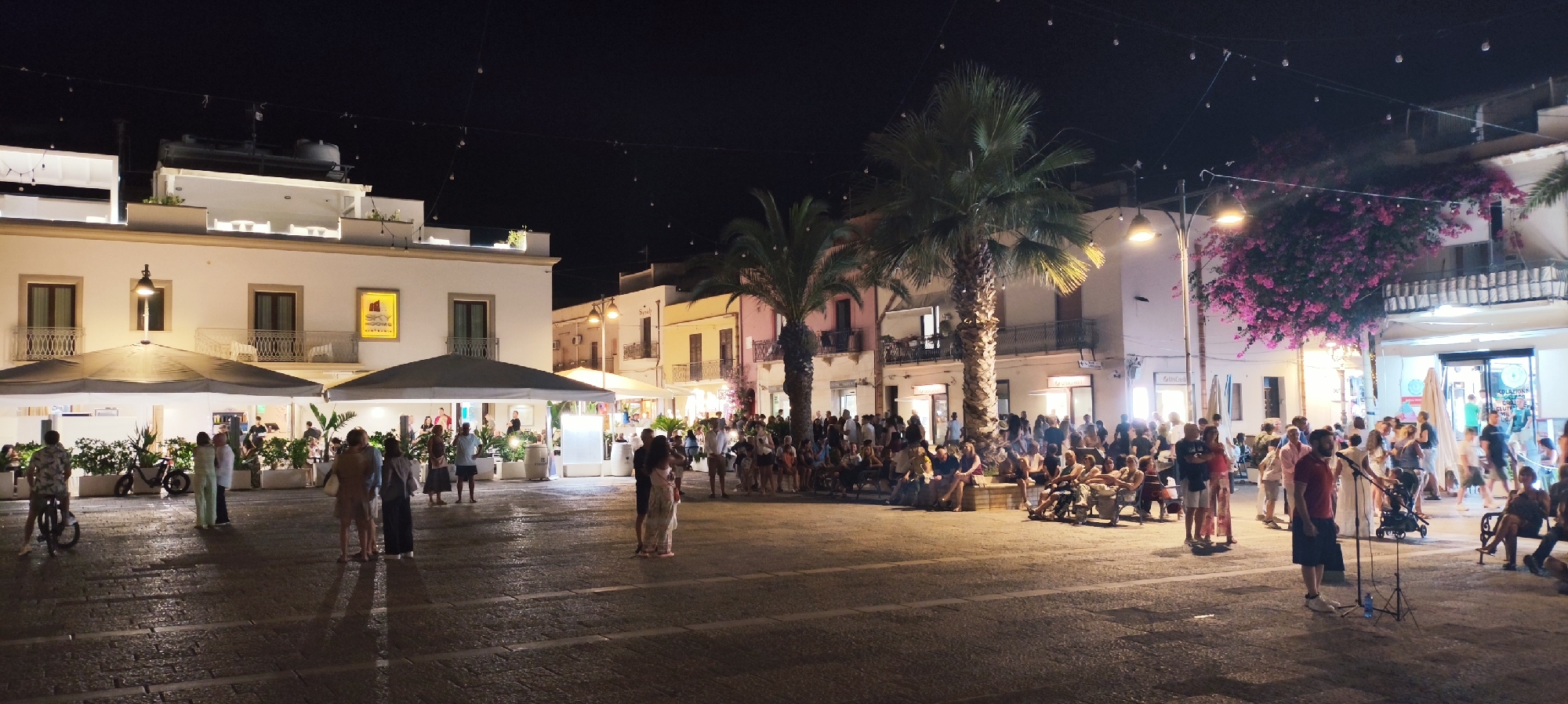 The image depicts a lively evening scene in the piazza of San Vito Lo Capo, Italy. The piazza is paved with cobblestones and is illuminated by string lights and building lights, creating a warm and inviting atmosphere. On the left, a white building with multiple stories features balconies and large umbrellas providing shade for outdoor seating areas. In the background, several buildings with colorful facades, palm trees, and vibrant pink bougainvillea add to the Mediterranean charm. A crowd of people is gathered in the piazza, some sitting and conversing, while others stand and observe. In the foreground, a man stands in front of a microphone on a tripod, suggesting a performance or event. People are casually dressed, and the overall mood appears relaxed and social.