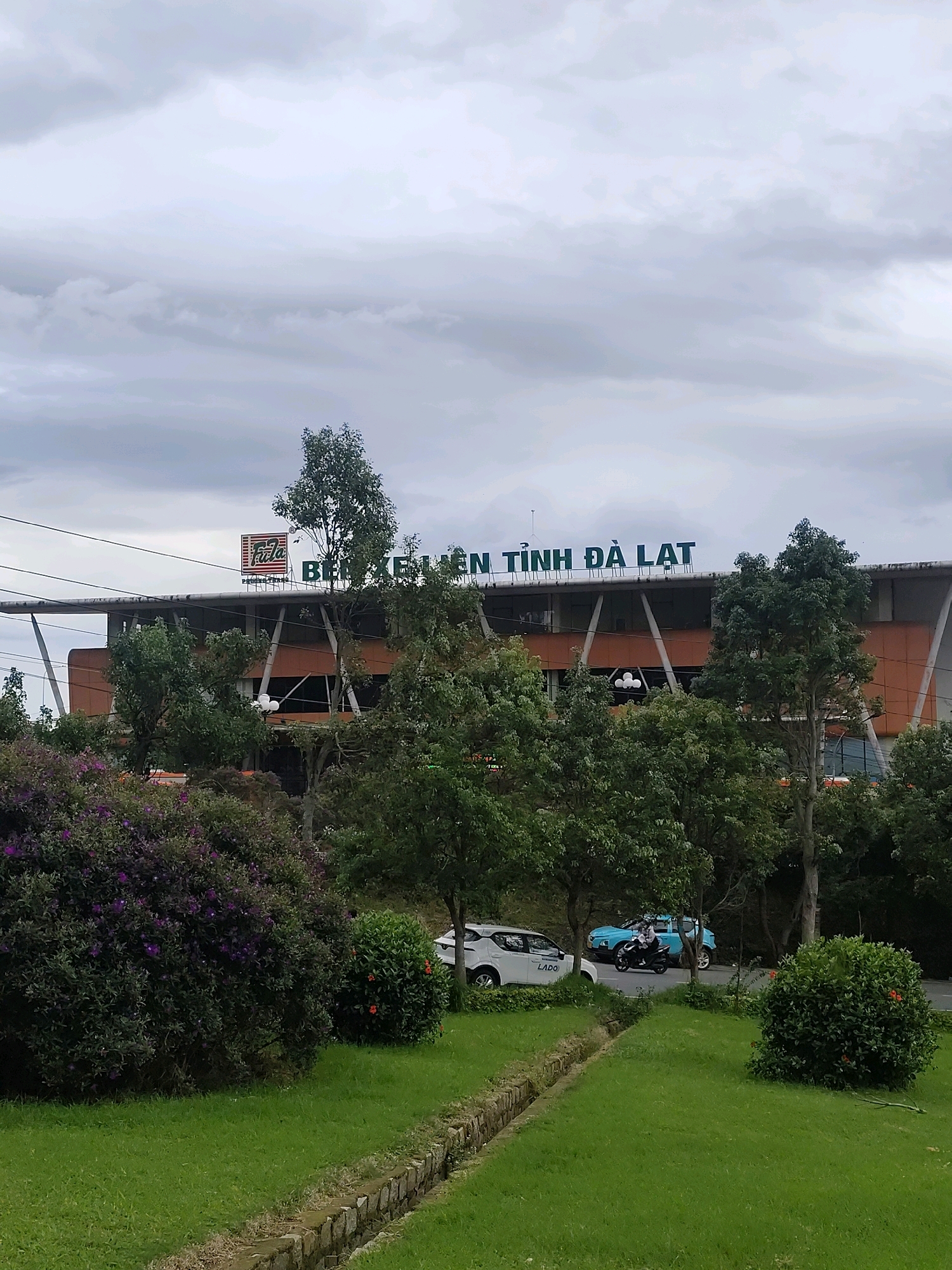 The image captures a cloudy day scene in Da Lat, Vietnam, featuring a modern building with large windows and an orange facade, topped with green lettering that reads "BEN XUAN TOEN DA LAT". A prominent logo, a red square with "FFLA" and "PHUNG TXEO" below it, is visible on the left side of the building. In the foreground, a well-maintained grassy lawn is bordered by a stone-lined ditch. A lush bush with purple flowers stands to the left, while a smaller green bush with red flowers is situated to the right. A road runs behind the building, where a white car, a blue car, and a person on a motorbike are present. Trees surround the scene, contributing to the natural ambiance.
