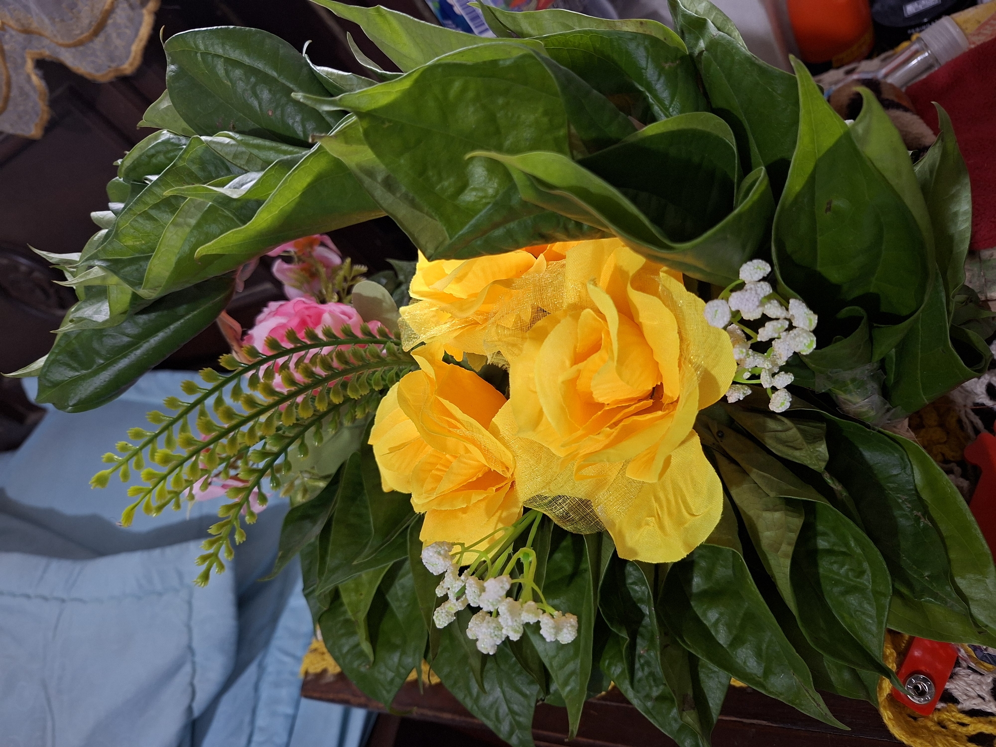 A vibrant bouquet of artificial flowers and foliage is prominently displayed in an indoor setting in Lhokseumawe, Indonesia. The central elements are three large, bright yellow roses, crafted from fabric and mesh, with a soft, textured appearance. These are complemented by small clusters of white artificial flowers, reminiscent of baby's breath, interspersed among the larger blooms. A hint of pink artificial flowers is also visible in the upper left background.
The flowers are set against a backdrop of numerous large, glossy dark green leaves, which strongly resemble betel leaves (Piper betle), traditionally significant in Indonesian culture. A delicate branch of light green, fern-like artificial foliage extends from the left side of the arrangement, adding varied texture.
The bouquet appears to be resting on a surface, likely a table, as suggested by the background elements. To the left, a light blue fabric, possibly a curtain or tablecloth, is visible, along with dark wooden furniture and a glimpse of an ornate, possibly gilded, decorative pattern. The right side of the background reveals fragmented views of household items, including an orange container, a piece of red fabric or strap with a silver snap button, and a section of yellow and white crocheted or woven material.
There is no specific activity depicted, but the arrangement suggests it serves as decoration for a celebration, event, or perhaps a cultural or religious ceremony, particularly given the traditional use of betel leaves in Indonesia. The lighting suggests well-lit indoor conditions, either during the day or under artificial light. No visible text is present in the image.