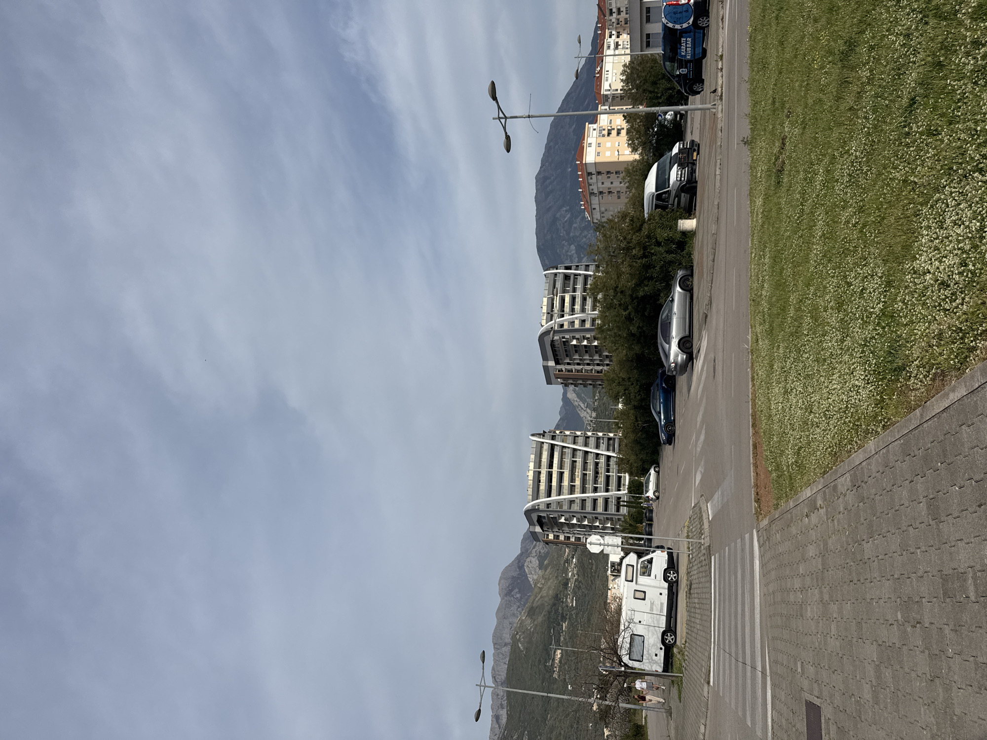 The image captures a street scene in Bar, Montenegro, featuring a row of parked vehicles along the right side of the frame, including a white camper, a dark blue car, a silver sedan, and a white van. In the background, modern residential buildings with curved balconies rise towards the mountains. A grassy area with small white flowers borders the parking. A sidewalk with a crosswalk is in the foreground. Street lights are intermittently placed along the road. The sky is overcast with light clouds. The landscape showcases a mix of urban development against a backdrop of natural mountainous terrain.