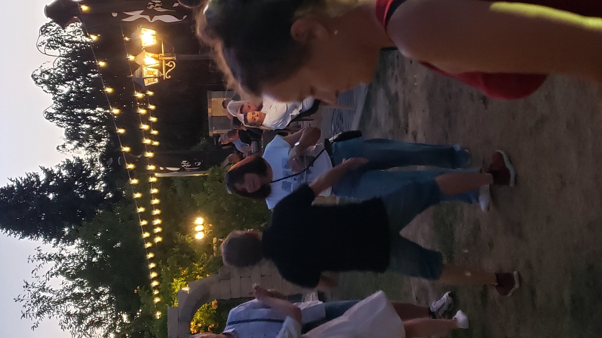 The image depicts an outdoor evening gathering, possibly at a venue in Prešov, Slovakia, featuring string lights and ambient lighting from a lamppost. In the foreground, two individuals are visible dancing. A person in a black t-shirt and denim shorts is facing away from the camera, while a woman in a white t-shirt and jeans is facing towards them, appearing to be dancing as well. Behind them, other people are seated at tables and standing around. The environment includes trees and what appears to be a stone structure or archway on the left. The overall atmosphere suggests a relaxed social event.