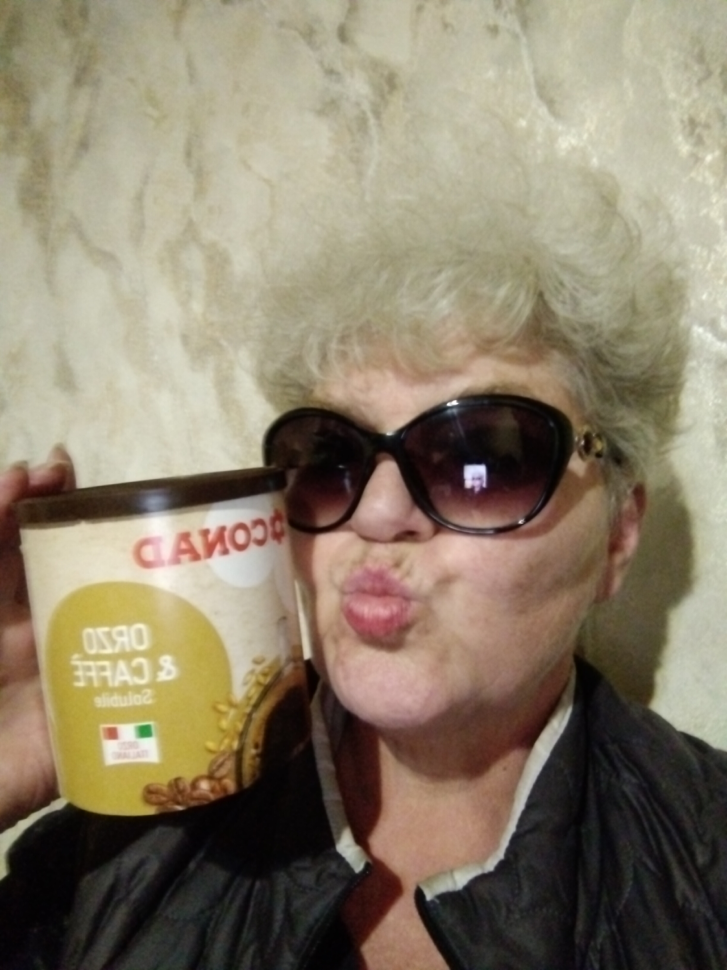 An older woman with voluminous, curly grey hair is featured in an indoor, close-up shot. She is wearing large, dark cat-eye sunglasses and a dark quilted jacket with a light-colored collar lining. Her face is turned slightly towards the viewer, and she has a playful expression with puckered lips.
She is holding up a light-colored, cylindrical container of what appears to be a beverage or food product. The brand name "CONAD" is visible in red text, appearing mirrored/backward on the container. Below it, the mirrored text "ORZO E CAFFE Solubile" (barley and coffee soluble) and a mirrored Italian flag symbol are discernible. The container also features graphics of coffee beans and possibly barley or wheat grains.
The background is a light-colored, textured wall, possibly stucco or wallpaper. The lighting suggests an indoor setting, likely with artificial light. The woman's expression conveys a sense of playfulness or lightheartedness. The image provides no specific cues for time of day or weather, but the location context is given as Horishni Plavni, Ukraine.