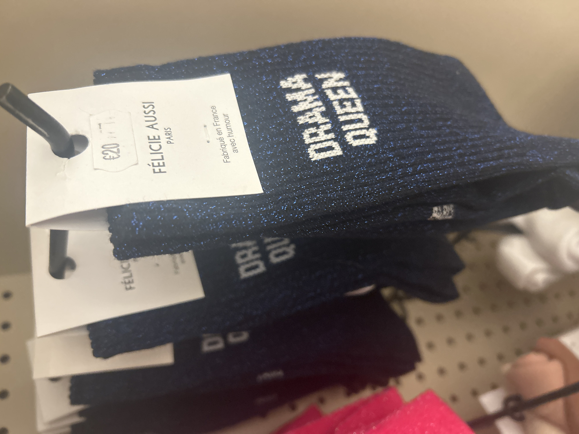 The image displays a retail store setting, focusing on several pairs of socks hanging on a black metal display hook. The most prominent pair of socks, positioned in the foreground, is dark blue with a subtle metallic glitter effect. White, block-letter text reading "DRAMA QUEEN" is embroidered vertically on the side of this sock. A white paper tag is attached to the foremost pair of socks, displaying a price of "€20.77" within an oval outline at the top. Below the price, the brand name "FÉLICIE AUSSI" is printed in bold capital letters, followed by "PARIS" underneath, indicating the brand's origin or association with the city of Paris, France. Further text on the tag reads "Fabriqué en France avec humour" (Made in France with humor). Behind the leading socks, other pairs, primarily dark blue, are visible, along with a partially obscured bright pink item on the far left. The background is a blurry beige or light-colored wall, possibly a pegboard display, showing circular indentations. No individuals are visible in the frame. The presence of "PARIS" on the product tag directly associates the brand and potentially the retail location with the city of Paris, France.