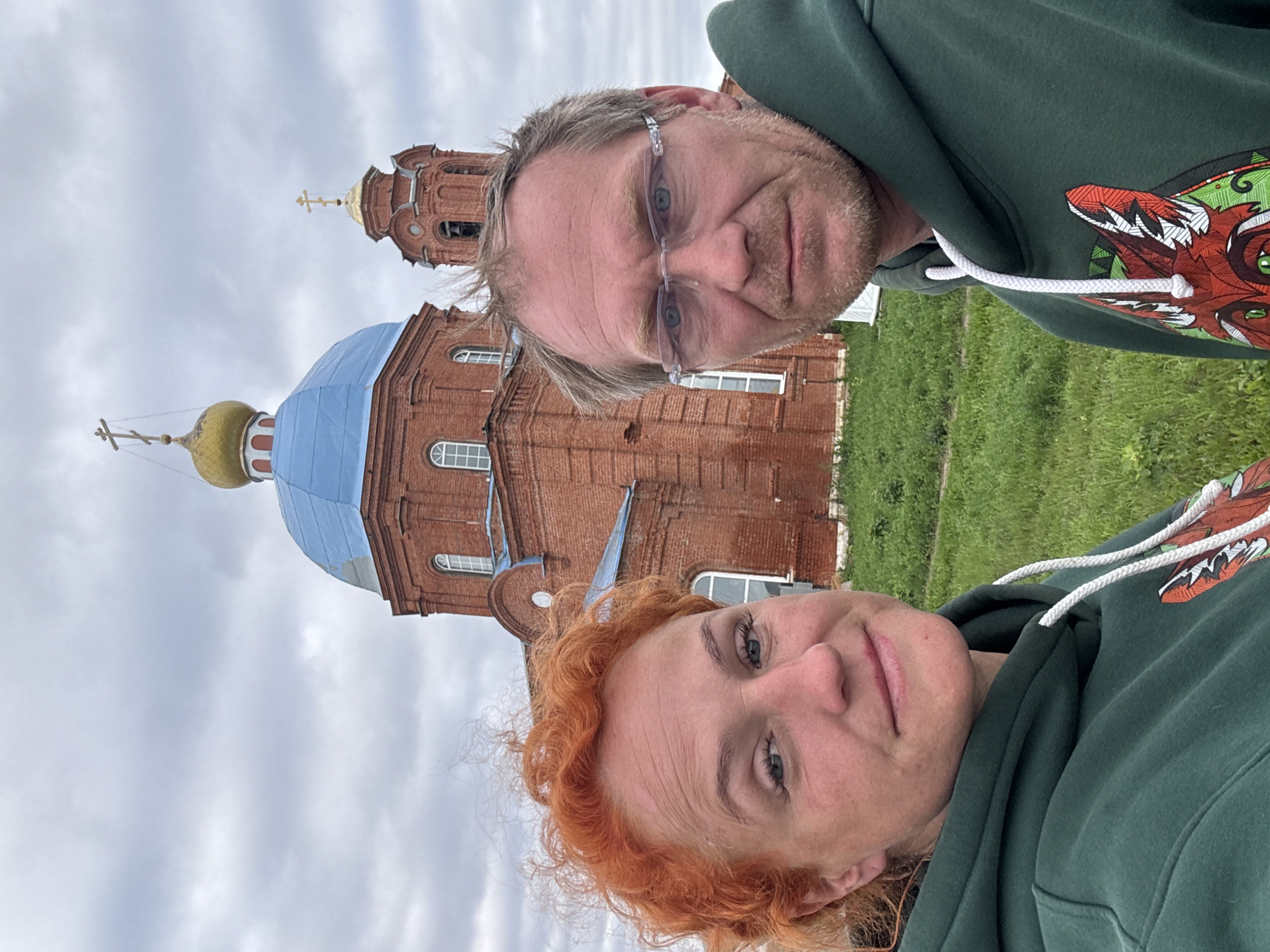 This is a selfie taken outdoors in front of a red brick church with a blue dome and a gold cupola. A woman with bright red, curly hair is in the foreground on the left, and a man with short, light brown hair and glasses is on the right. Both are wearing dark green hooded sweatshirts. The church is in the background. The sky is overcast. The image appears to be taken in or near the village of Anatesh, Russia.
