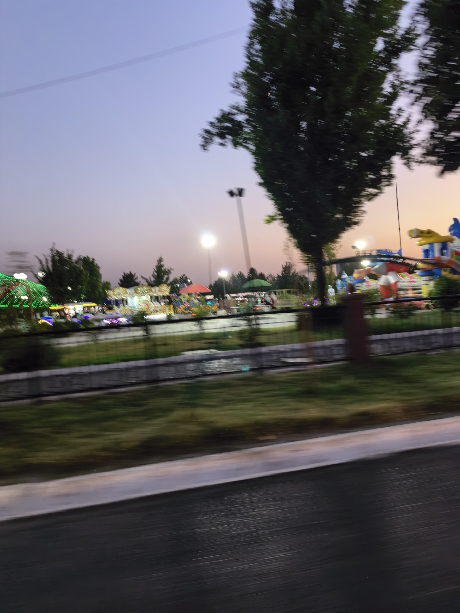 The image depicts a blurry amusement park in Fergana City, Uzbekistan, at dusk. The sky is a gradient of purple and pink, transitioning to a lighter blue. In the foreground, a blurred asphalt road is visible. Beyond this, a strip of grass is present, separated from the park by a low concrete barrier. The park itself features various rides and attractions, illuminated by colorful lights, including a Ferris wheel, a carousel, and what appears to be a dragon-themed ride. Trees are scattered throughout the park. Streetlights are also visible, emitting a soft glow. The overall impression is that of a lively entertainment venue captured in motion, with the blur indicating a moving camera or vehicle.