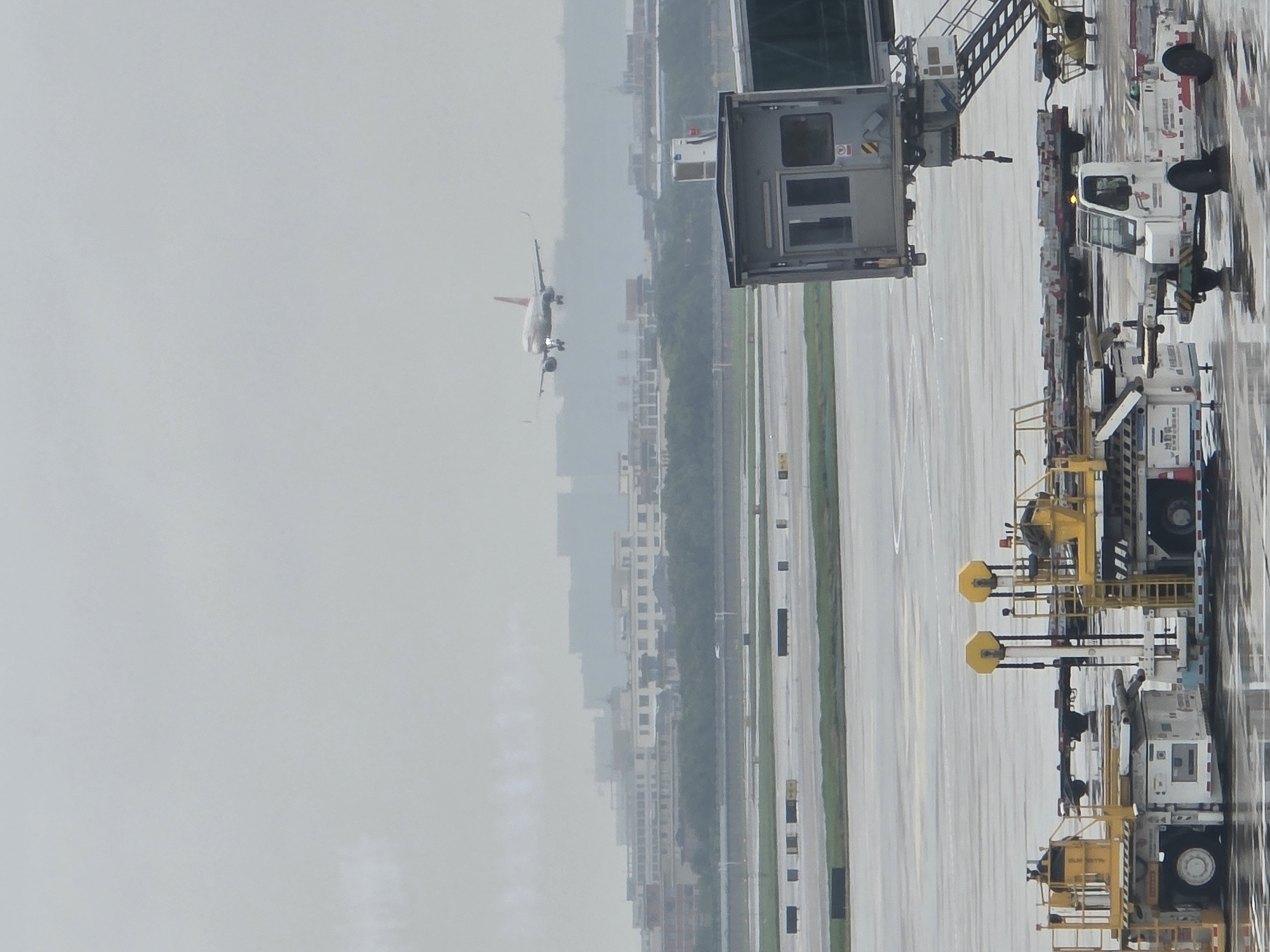 This image depicts an airport scene in Xiaoshan District, China, under a hazy sky. In the foreground, two yellow and white airport service vehicles are parked on the wet tarmac. One vehicle, on the right, is a white tow tractor with red accents, and another, on the left, is a larger white and yellow vehicle with a raised platform and wheels. A jet bridge extends from a building on the right side of the image towards the runway. In the middle ground, the wet tarmac of the airport runway is visible with various markings and some scattered black rectangular objects. In the background, a city skyline with low-rise buildings and trees can be seen through the atmospheric haze. An airplane, with its landing gear down, is descending towards the runway in the upper part of the image. The overall atmosphere is overcast, with wet surfaces suggesting recent rain. There are no people clearly visible in the image.