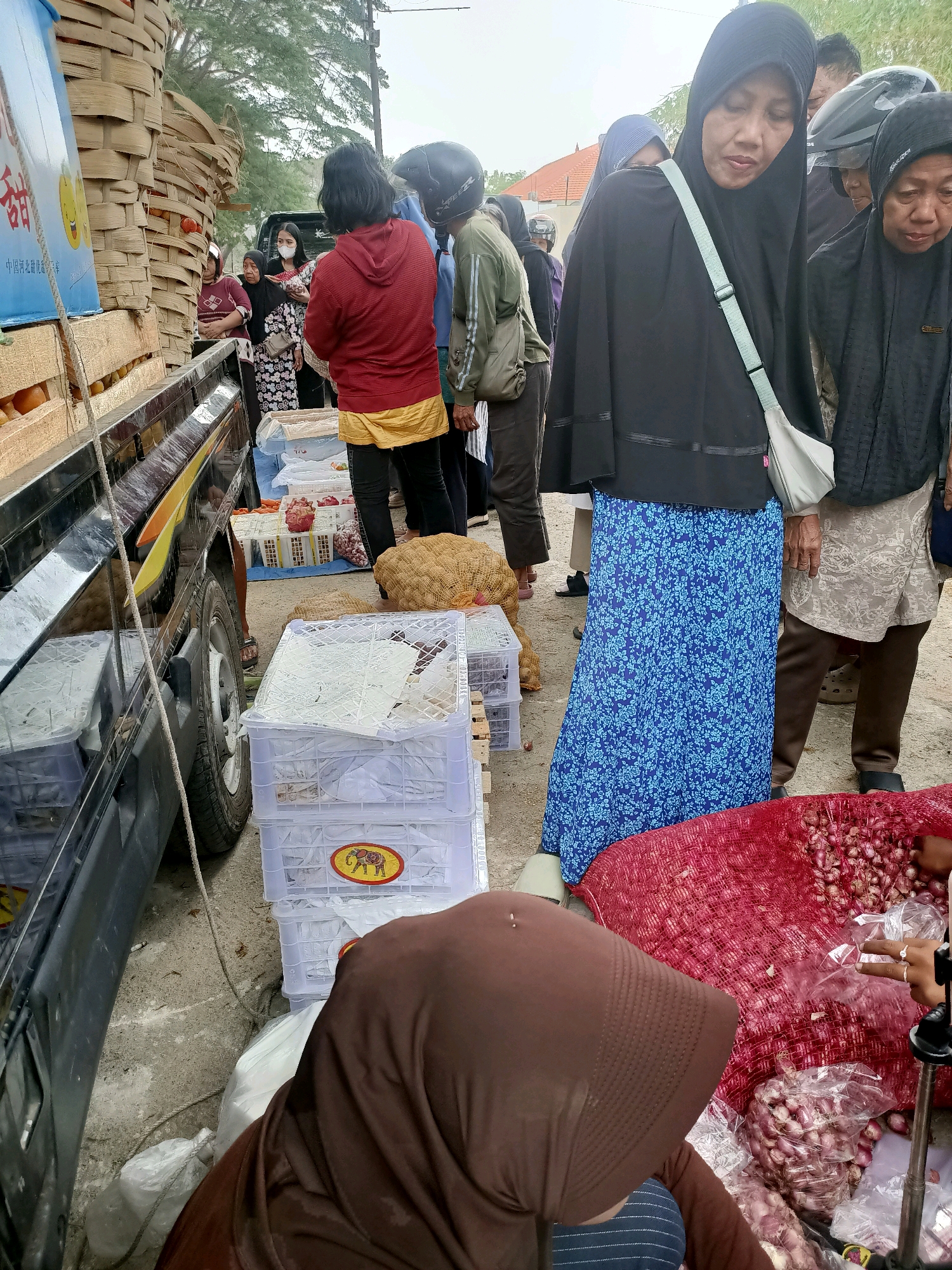 In Madiun, Indonesia, a group of people are gathered at an outdoor market, likely a wholesale produce market, unloading or arranging goods from a truck. The scene is characterized by an abundance of agricultural products, with large woven baskets filled with fruit, likely oranges, stacked high on the truck. Below the baskets, crates and sacks are visible, containing more produce such as onions, which are prominently displayed in large red mesh bags.
Several individuals are interacting with the goods. A woman in a black hijab and a blue floral skirt is looking down towards the produce, possibly selecting items or overseeing the unloading. Another woman in a red jacket and yellow skirt is standing near the truck, and a woman in a brown hijab is partially visible in the foreground, appearing to be tending to the onions. Other people in the background, some wearing hijabs and others not, are also present, suggesting a busy market environment. The ground is paved, and the lighting indicates it is daytime, possibly overcast. The overall impression is one of commerce and the distribution of fresh produce in a bustling local market.
